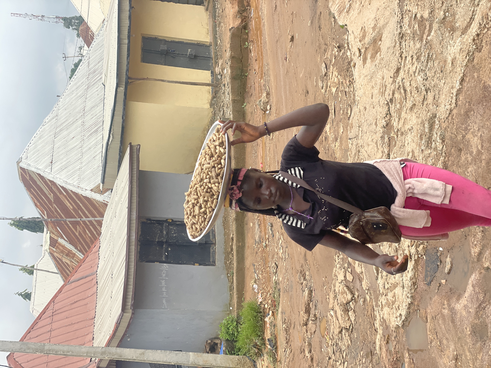 A young woman walks on a dirt road in Kurunduma, Nigeria, carrying a large metal platter of peanuts on her head. She wears a black t-shirt with a white striped collar, pink leggings, and a light pink sweater tied around her waist. A small brown crossbody bag hangs from her shoulder. The ground is a mixture of mud and rocky debris, with several puddles of water visible. In the background, several buildings with corrugated metal roofs are visible, some appearing weathered. The sky is overcast.