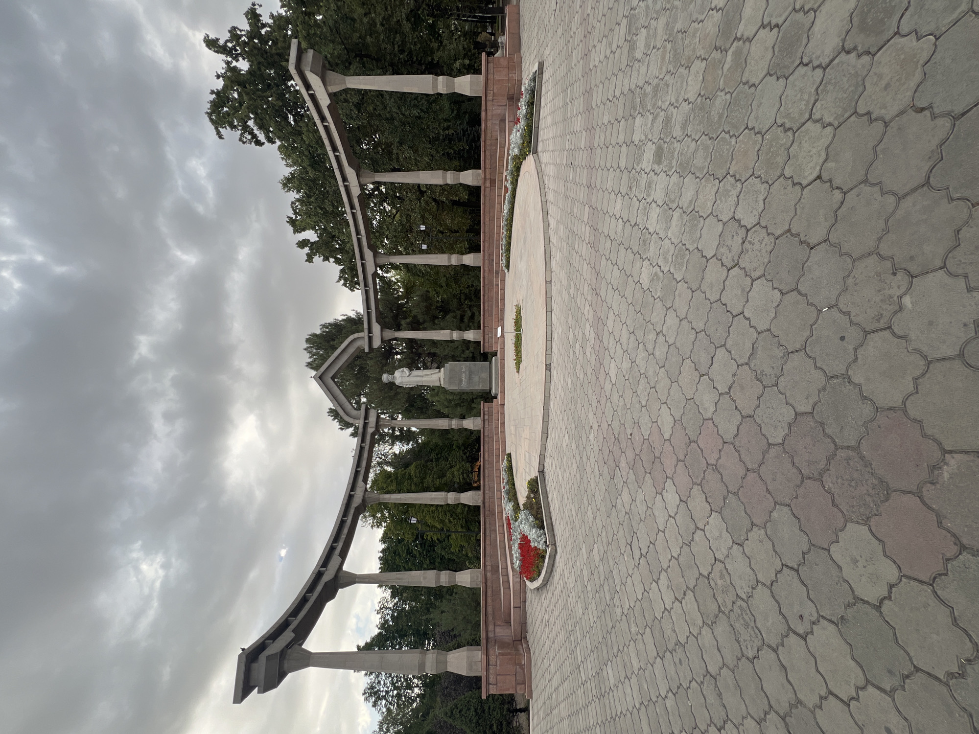 The image shows a memorial complex with a statue in Bishkek, Kyrgyzstan. A paved plaza leads to a raised circular platform with a statue of a man in traditional clothing. The statue stands on a pedestal with an inscription. Flower beds with red and white flowers surround the platform. A curved, modern pergola made of concrete pillars and beams arches over the plaza. The sky is overcast with heavy clouds. The background features lush green trees.