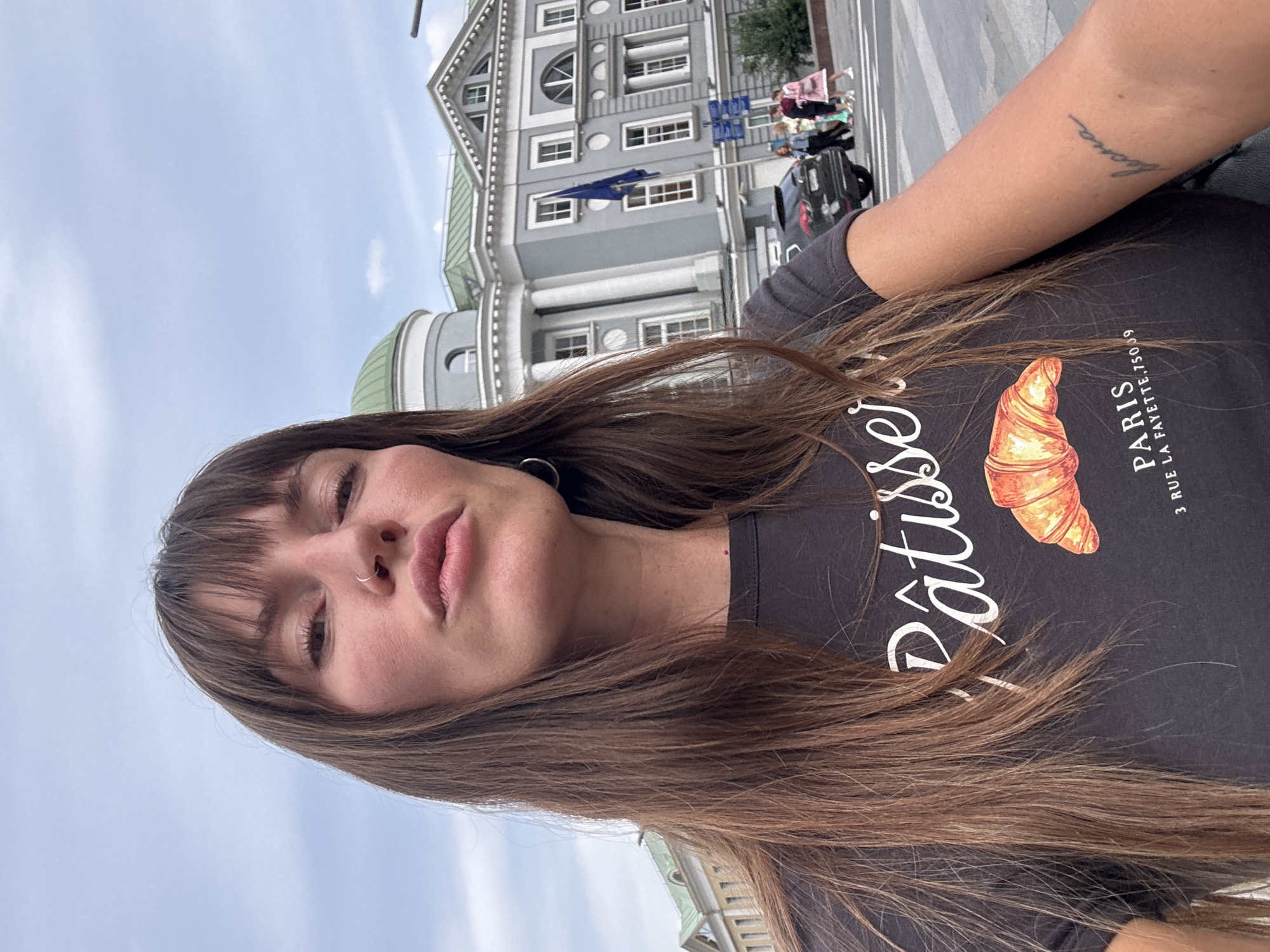 A young woman with long brown hair and bangs poses for a selfie in front of a building with classical architecture. She is wearing a dark t-shirt with a croissant graphic and the words "Pâtissier" and "PARIS" printed on it. A tattoo of the word "весна" (spring in Russian) is visible on her left forearm. In the background, a large grey building with a green dome and a blue flag can be seen. Several people and a black car are crossing a street in front of the building. This location is the main building of the State Historical Museum on Red Square, Moscow, Russia.