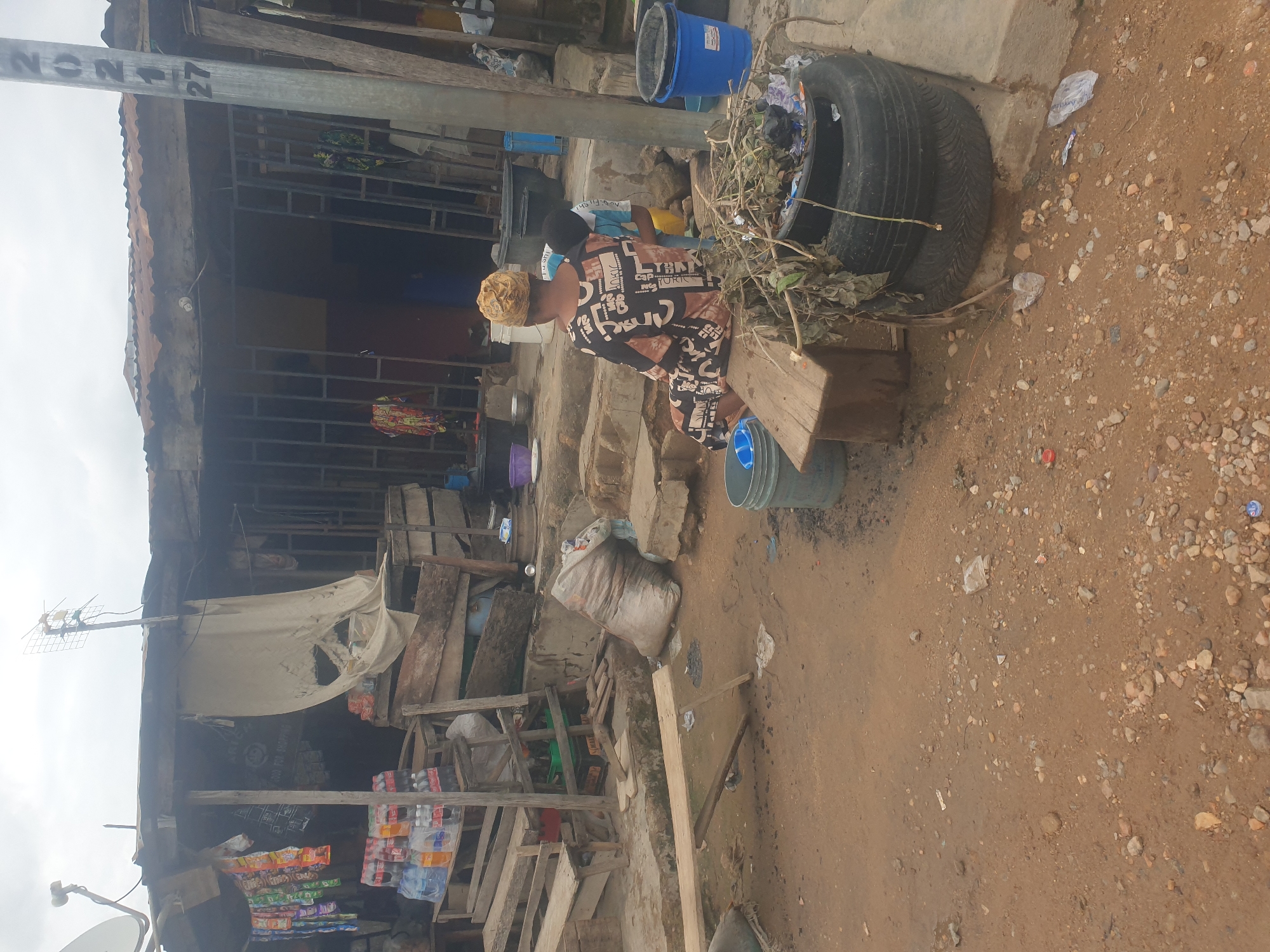 The image shows an outdoor scene in what appears to be a developing or impoverished area. A woman is seated on a wooden structure near a tire filled with debris, possibly burning or processing waste. The surroundings include dilapidated buildings constructed from wood and metal, displaying signs of disrepair and improvisation. The foreground is a dirt and gravel ground with scattered debris. Overhead utility lines and poles are visible. Some goods such as drinks and snacks are displayed on the front of one of the buildings. There is a blue bucket in the image.