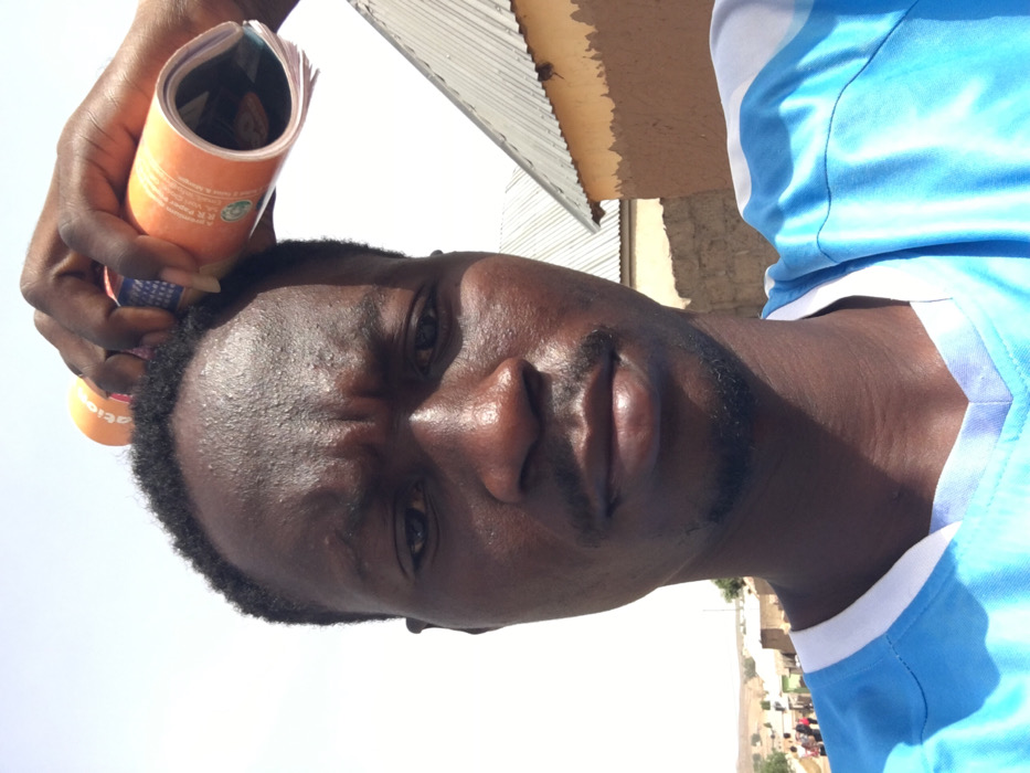 This image, captured in Pindiga, Nigeria, presents a close-up portrait of an adult male. The subject has dark skin, short black hair, a thin mustache, and a chin beard. He wears a light blue t-shirt with a white collar and light blue/white patterned shoulders. His right hand is positioned above his head, holding a rolled-up orange magazine, which appears to be utilized as a sunshade. Text, partially visible on the magazine, includes "...ation". His expression is neutral, with a slightly furrowed brow, and he looks directly into the camera. The background features a bright, clear sky indicative of strong daylight conditions. To the right of the subject, a building with visible mud-colored walls and a corrugated metal roof is present. Further in the distance, a blurry street or path contains several indistinct individuals and additional low-rise structures, consistent with a populated, semi-arid urban environment.