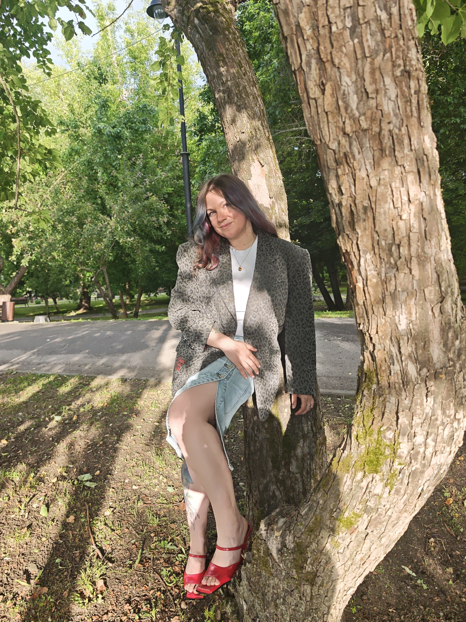 A young woman with dyed hair is sitting on a tree trunk in a park. She is wearing a leopard print blazer over a white top and a denim skirt. Red heeled sandals are on her feet. The scene is set in a park in Dubna, Russia, with trees and greenery in the background, suggesting it is daytime during warm weather. The lighting indicates a sunny day, with dappled sunlight on the ground. The woman is looking towards the camera with a slight smile, conveying a relaxed and confident mood. There are no visible text elements in the image.