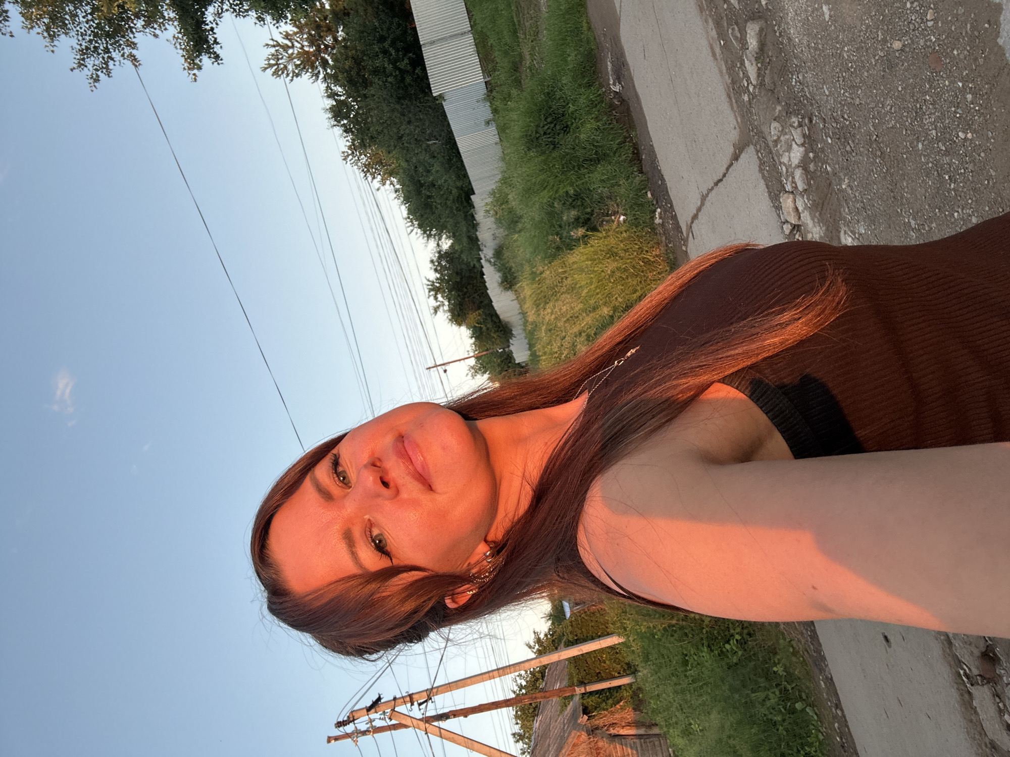 A woman with shoulder-length brown hair and green eyes is photographed outdoors in a selfie at sunset. She is wearing a dark brown ribbed tank top and a silver necklace. The warm golden light of the setting sun illuminates her face and right arm. In the background, there are power lines and poles, a rustic wooden building, tall green grass, a corrugated metal fence, and a paved road with scattered rocks. The sky is a clear, light blue with a few wispy clouds. The location appears to be a rural or semi-rural area on the outskirts of the city of Chaikovsky, Russia.