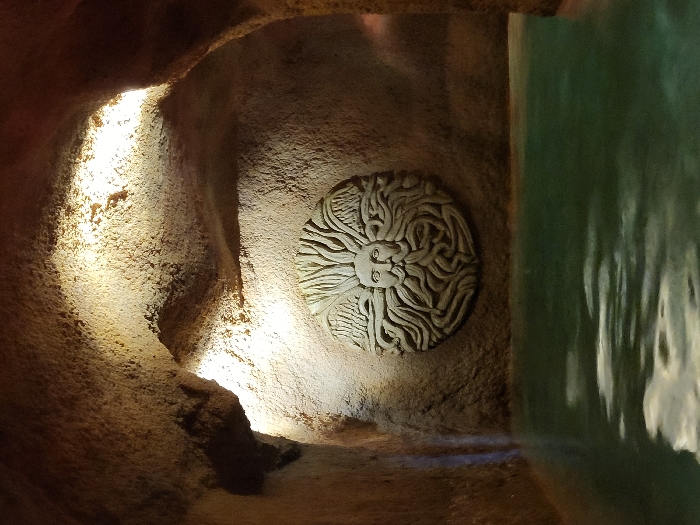 A textured, circular sun sculpture adorns a wall within a cave-like setting. Light illuminates the wall around the sculpture from the left side. To the right, a glass structure allows light to shine through and creates a blurry, greenish image. The cave walls and floor are a mix of sandy and brown hues. The scene appears to be indoors.