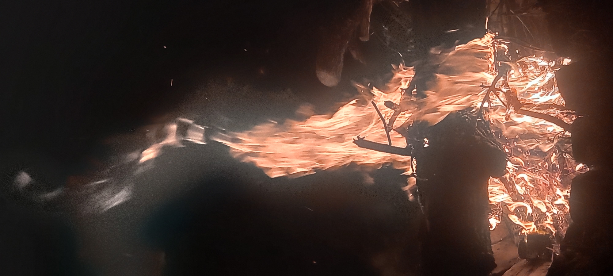 This vertical image captures a large, active bonfire burning in darkness in the city of Kryvyi Rih, Ukraine. The fire dominates the lower and central portions of the frame, characterized by intense orange and yellow flames that extend upwards in dynamic, swirling columns. Lighter, semi-transparent smoke plumes are visible above the main fire, illuminated by the underlying heat and mixing with the ascending flames. Scattered bright sparks are discernable within the smoke and against the dark background, indicating small burning particles being carried aloft.
The base of the bonfire consists of various wooden materials, including several substantial logs and numerous smaller branches and brush, all actively consumed by the fire. A prominent, thick piece of broken wood is visible in the mid-section of the main flame column. To the right, partially obscured by the intense glow, a darker, elongated object, consistent with another log, is positioned within the fire's periphery. The immediate environment surrounding the bonfire and the entire background are uniformly dark, indicating a nighttime setting with the fire as the sole significant light source. No individuals or additional objects are explicitly identifiable in the scene.