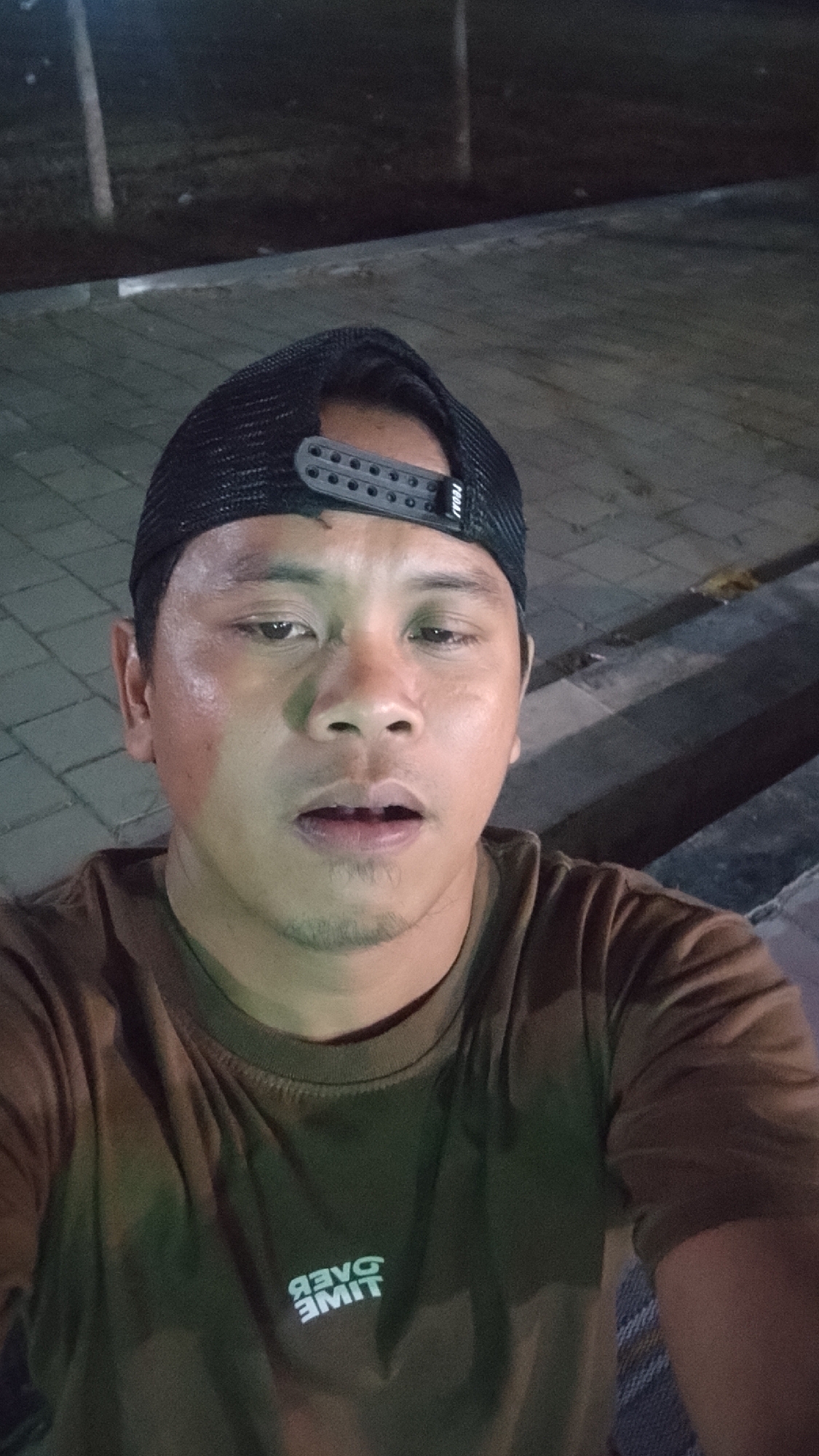 A man is taking a selfie outdoors at night. He is wearing a brown t-shirt with white lettering on the front, a black trucker hat, and a black baseball cap with adjustable snapback closure. The man's face is the primary focus of the image. He has short dark hair, a fair complexion, and is looking directly into the camera. His mouth is slightly open. The background shows a paved area with square tiles, and some blurred trees and a railing. The location is identified as Karanglangu, Indonesia.