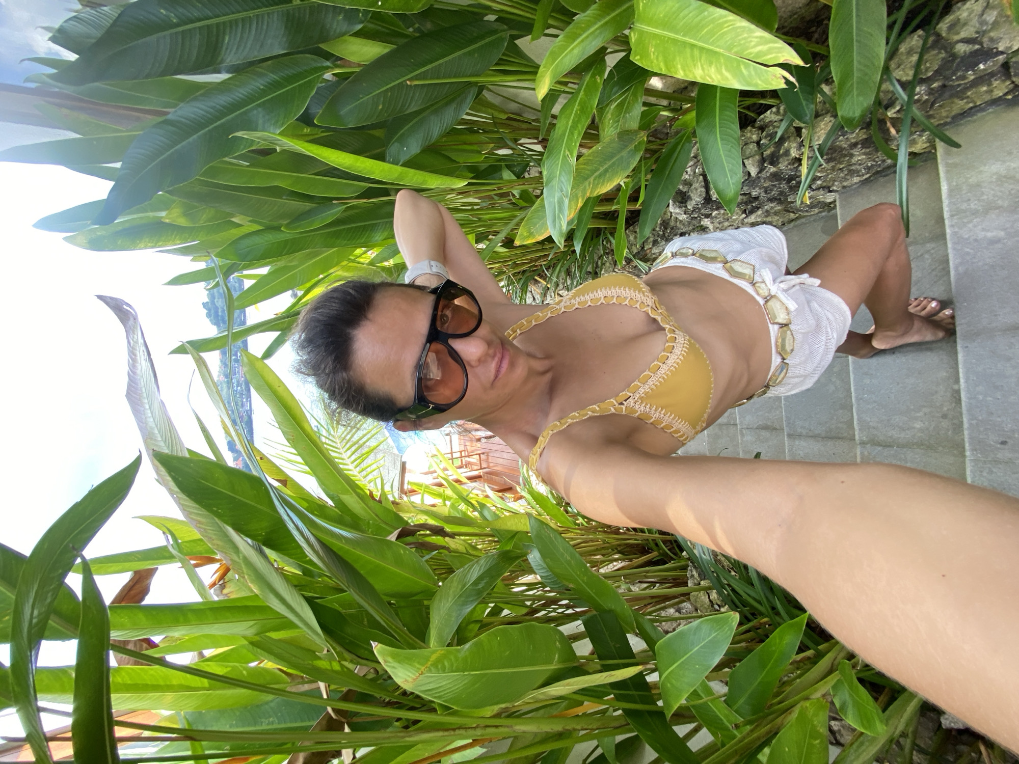 A woman with dark hair and sunglasses poses for a selfie on a stairway lined with lush green tropical foliage. She wears a yellow bikini top and white shorts with a decorative belt. In the background, a body of water and some buildings are visible, suggesting a coastal location. The scene is outdoors, under bright daylight.