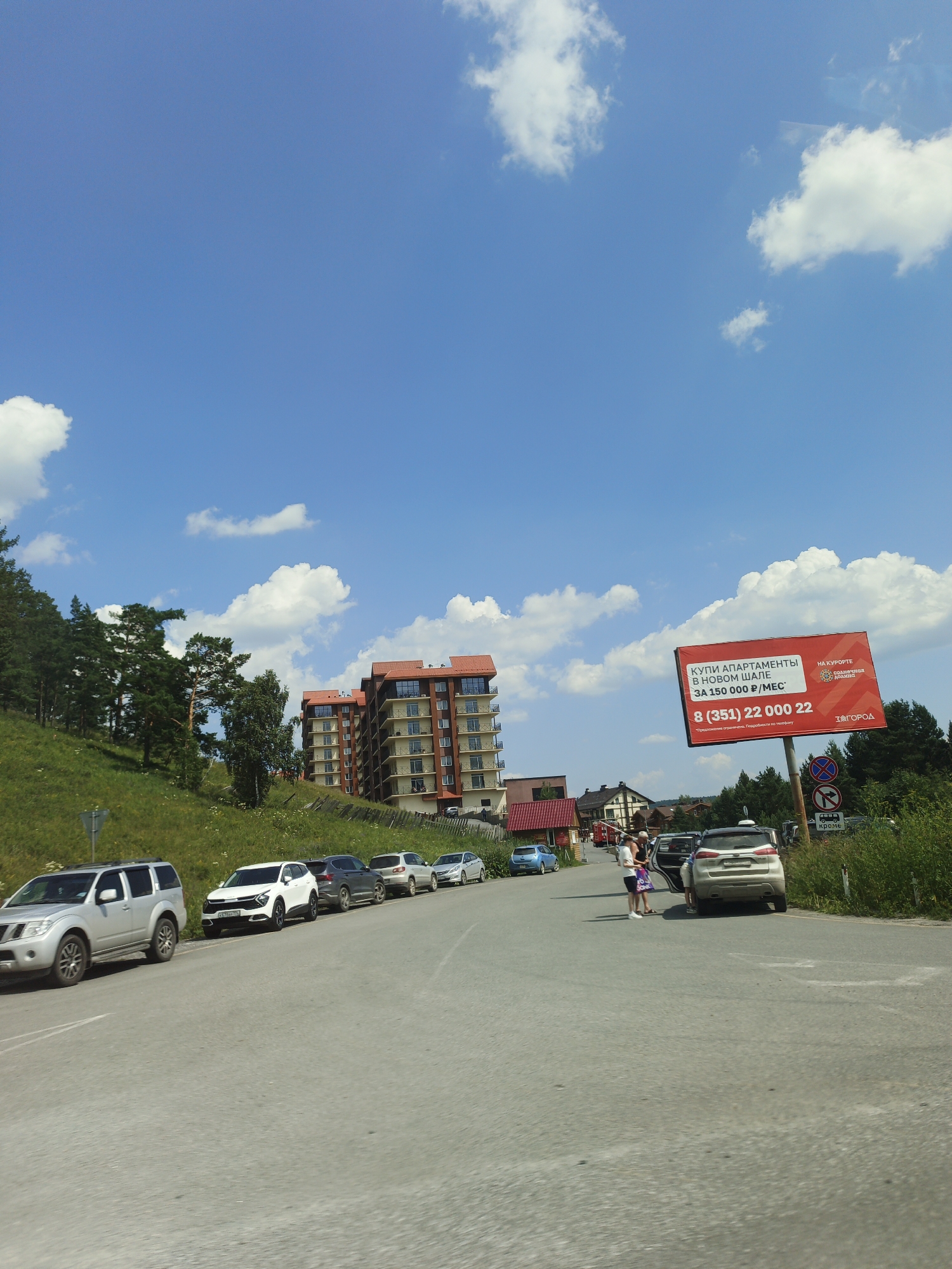 The image captures a street scene in the city of Syrostan, Russia, under a bright blue sky dotted with white clouds. On the left side of the road, a green, grassy hill rises, sparsely populated with coniferous trees. Several multi-story buildings with reddish-brown roofs and balconies are visible on the hillside, indicating a residential or resort area. A billboard advertising apartment sales in "Novom Shalle" for 150,000 rubles per month, along with a phone number and "Na Kurorte Solnechnaya Polyana" (At the Solnechnaya Polyana Resort), is prominently displayed on the right side of the road. The road itself is paved and curves slightly to the right. A line of cars is parked along the right edge of the road. In the middle ground, two individuals stand near an open car door. To the far right, lush green foliage borders the road, and several traffic signs are visible, including one indicating a restriction on entry and another with the word "Кроме" (Except).