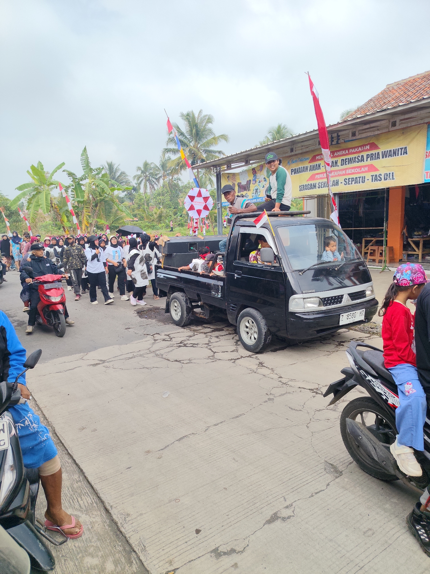 In Sukaratu, Indonesia, a parade is underway on a paved road under an overcast sky. A black pickup truck, adorned with Indonesian flags, is the central focus, carrying several people, including two young boys in the cab and a man and a young woman standing in the truck bed. The truck is followed by a procession of people walking, some wearing matching black and white outfits, and others in casual attire. A man on a red motorcycle leads the group, while another person is partially visible on a motorcycle in the foreground. Buildings line the right side of the road, one with a large banner that advertises clothing and school supplies. Palm trees and other vegetation are visible in the background, suggesting a rural or semi-urban setting. Red and white pennants are strung along the side of the road, adding to the festive atmosphere of the event.