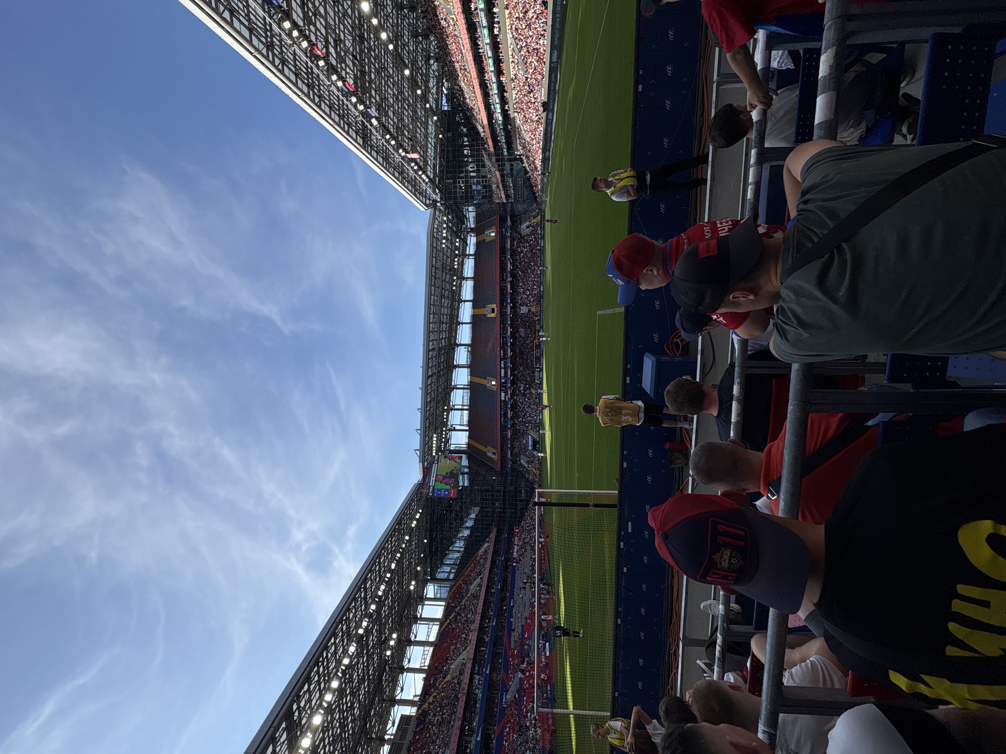 The image depicts a scene at a football stadium in Khimki, Russia, under a bright blue sky with scattered clouds. The foreground shows spectators seated in the stands, facing a lush green football pitch. In the middle ground, players are visible on the field, engaged in a match. The stadium features tiered seating with a distinctive roof structure covering a portion of the stands. A large video screen is positioned in the distance, displaying content related to the game. The overall atmosphere suggests an active sporting event with a sizable audience.