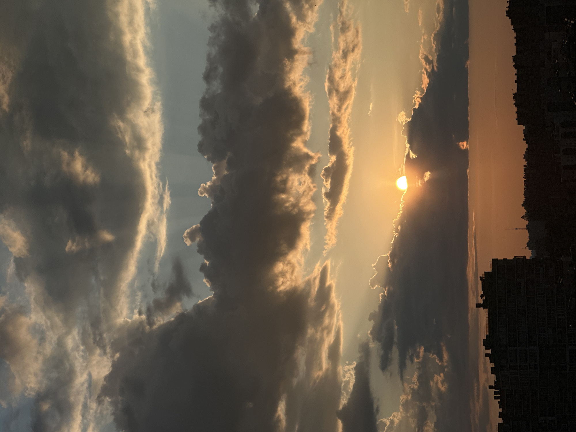 This is a vertical, outdoor, medium shot photograph of a sunset over a city. The sun is setting behind dark, dramatic clouds in the upper left quadrant of the image. Rays of sunlight are breaking through the clouds, casting a warm glow on the sky. Below the clouds, the horizon is lined with the dark silhouettes of buildings, including a large, modern apartment building in the lower left corner. The sky transitions from a bright orange near the horizon to a pale blue at the top of the frame. The scene captures a sense of twilight and the beauty of a cloudy sunset.