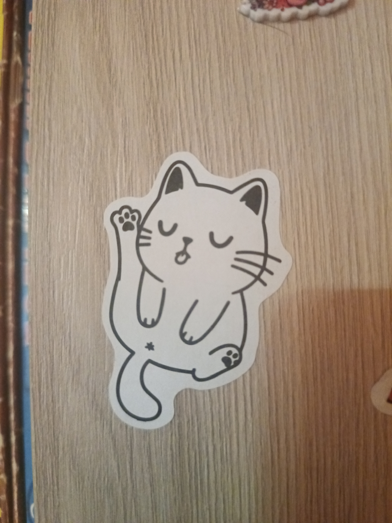 This media file is a photograph of a sticker of a cartoon cat. The cat is black and white with a simple line drawing style. It has closed eyes, a small mouth, and whiskers. One paw is raised in the air, and the other is resting on its lap. A tail curves around the body, and there is a small star symbol near the tail. The sticker is placed on a wooden surface that appears to be a wall or furniture. The wood grain is visible, and it has a light brown color. There are other stickers partially visible in the image, one in the top right corner that appears to be colorful and possibly of fruit, and another small one in the bottom right corner. The overall scene is casual and appears to be taken indoors, possibly in a room decorated with stickers. There is no discernible time of day or weather. The emotional tone of the sticker is playful and cute. There is no visible text in the image.
