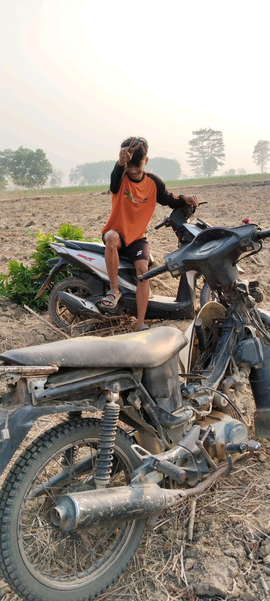 In Tanjung Ratu Ilir, Indonesia, a young person with dark hair and an orange and black long-sleeved shirt is sitting on a scooter in a dry, open field. The individual is making a peace sign with their right hand. The field is mostly dirt with some dry grass and scattered debris. Two scooters are visible in the foreground and midground. The scooter in the foreground is partially disassembled, with visible engine components and a rusty exhaust pipe. The scooter in the background is more intact, with a silver and white body. Trees and a hazy sky are visible in the distance, suggesting a rural or semi-rural setting.