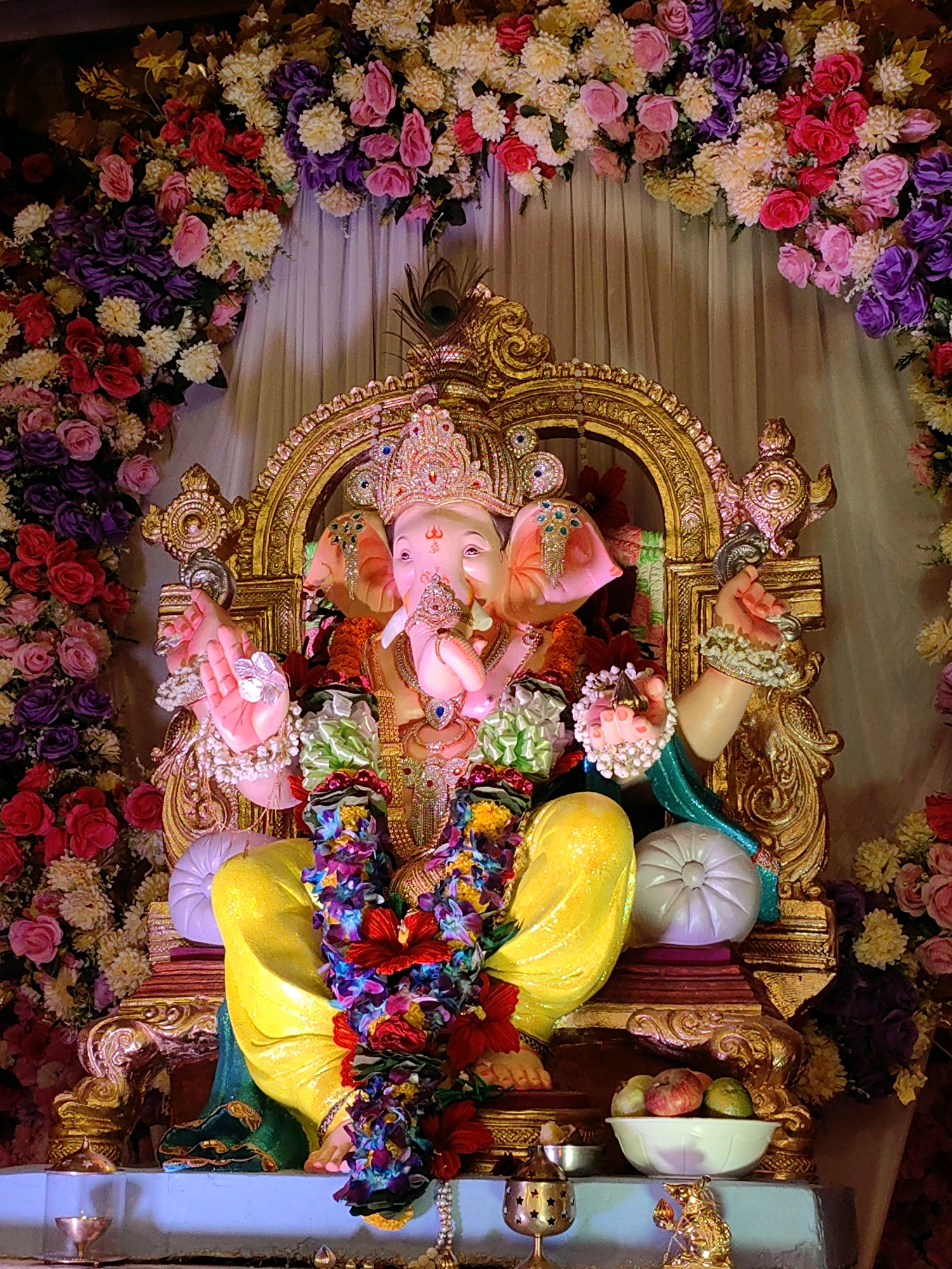The image shows a large statue of Ganesh, a Hindu deity, seated on a gilded throne. The statue is adorned with garlands of colorful flowers, beads, and intricate jewelry. Ganesh is depicted with his characteristic elephant head, a human body, and multiple arms, holding various symbolic objects. He is dressed in vibrant yellow attire.
The setting appears to be a temple or a home altar, decorated extensively with floral arrangements in shades of pink, red, purple, and cream. A backdrop of light-colored curtains is visible behind the statue. In the foreground, a small bowl of fruits, a decorative lamp, and a golden figurine of a mouse, Ganesh's vahana (vehicle), are placed on a platform. The overall impression is one of religious devotion and festivity, likely during Ganesh Chaturthi, a major Hindu festival celebrated in Mumbai, India. While the specific location within Mumbai is not identifiable from the image, the cultural context strongly suggests it is within the city during the festival period.