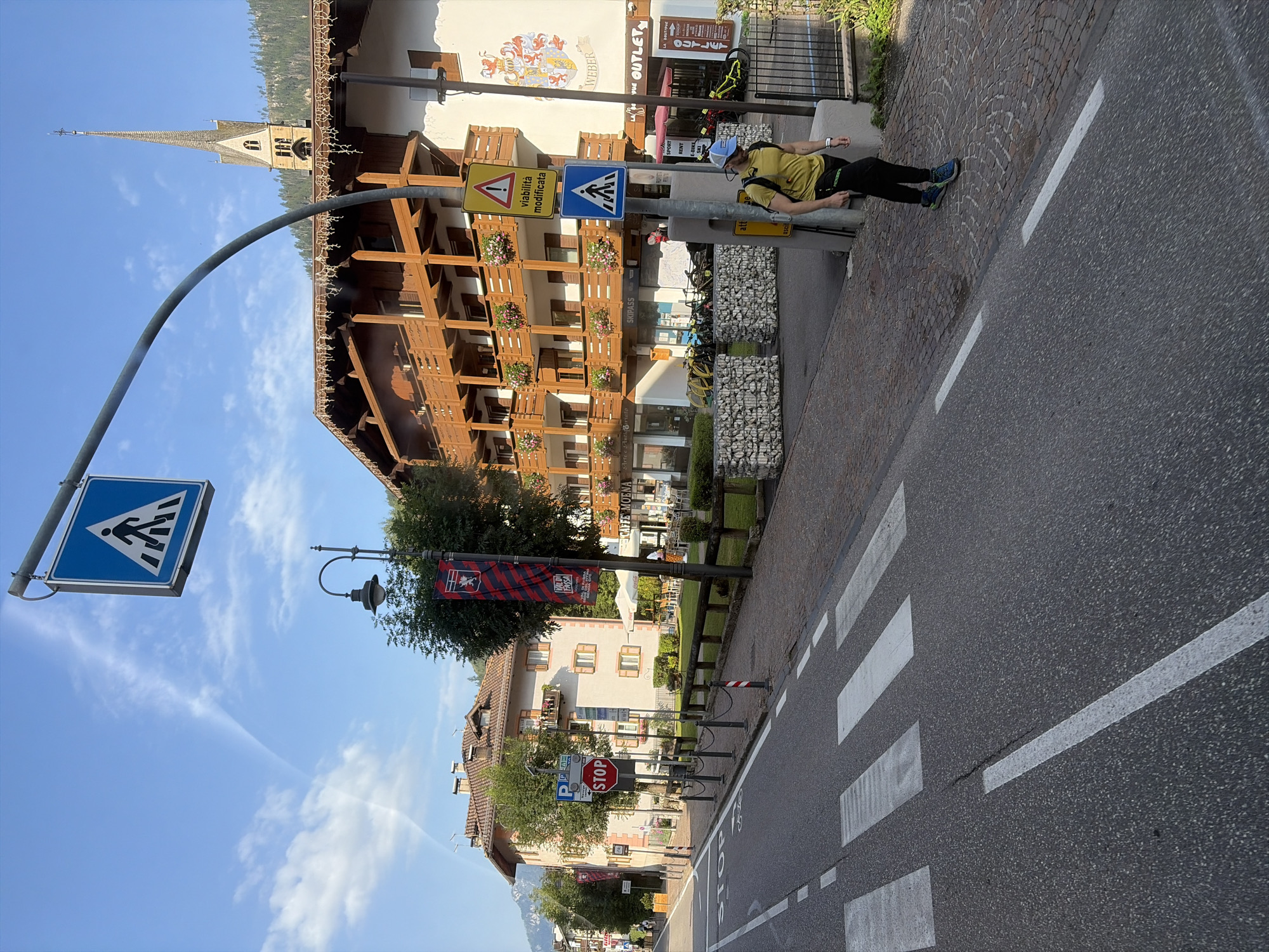 The image depicts a street scene in Moena, Italy, on a sunny day with a clear blue sky and scattered white clouds. In the foreground, a crosswalk is marked on an asphalt road, with white stripes indicating pedestrian passage. To the right of the crosswalk, a person wearing a yellow t-shirt, black pants, and blue and green athletic shoes is walking on a cobblestone sidewalk. The person is wearing a blue baseball cap and a backpack.
Further down the street, several buildings with wooden balconies and flower boxes line the sidewalk. One building has a tall, slender spire topped with a cross, suggesting it is a church. Various signs are visible, including a blue pedestrian crossing sign, a yellow sign with a warning symbol and text in Italian ("viabilità modificata," meaning "traffic modified"), and a red octagonal "STOP" sign. Banners with a crest and red and blue stripes hang from lampposts. In the background, trees and a mountain range are visible. The overall scene conveys a European town setting.