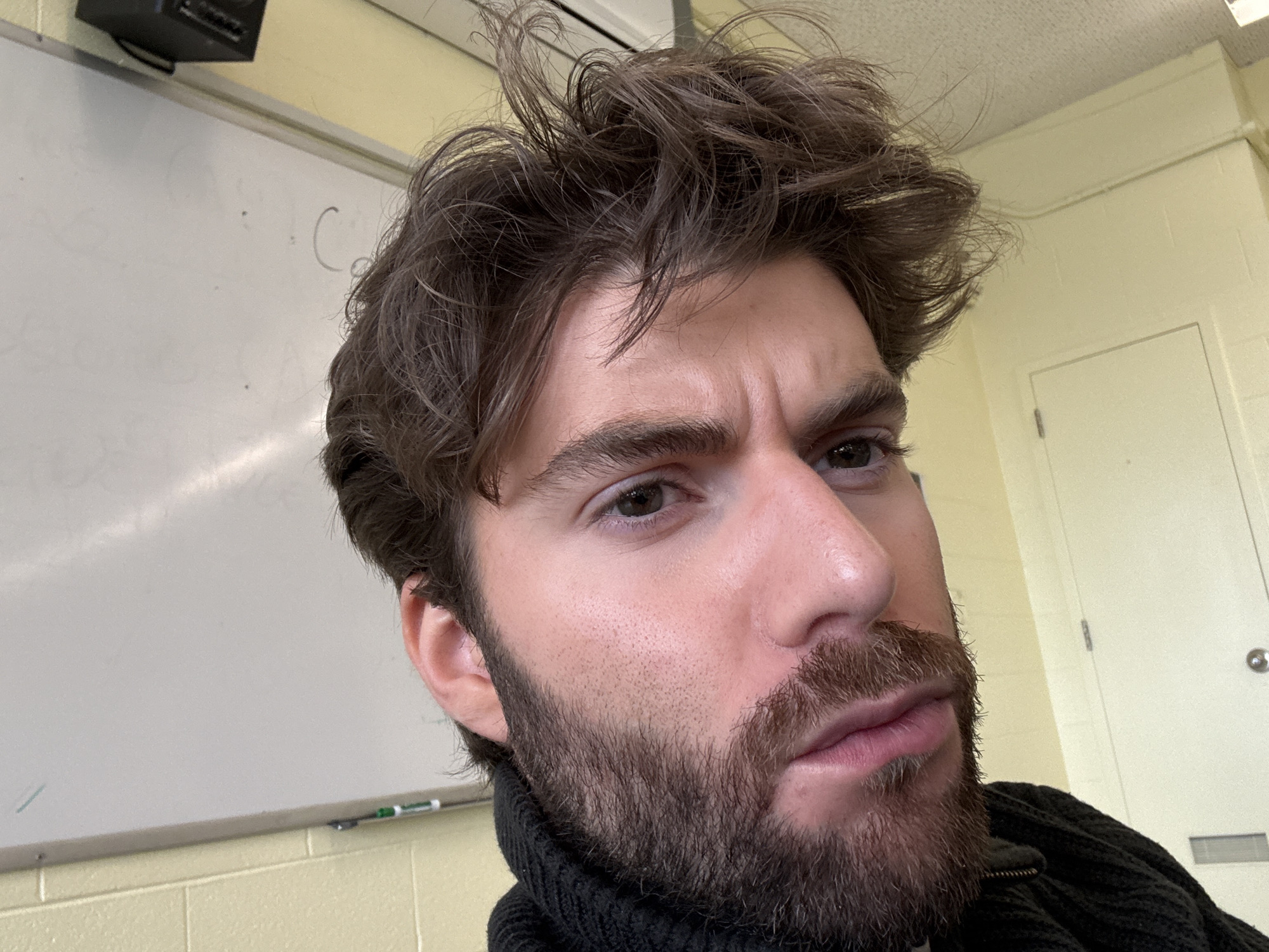 A close-up headshot shows a man with medium-brown hair, styled in a messy, tousled fashion. He has a thick brown beard. He is wearing a black turtleneck sweater. The background includes a white whiteboard with some writing on it and a yellow wall. A black speaker is mounted near the top-left corner. A door is visible on the right. The location in Montreal, Canada is not discernible from the image.