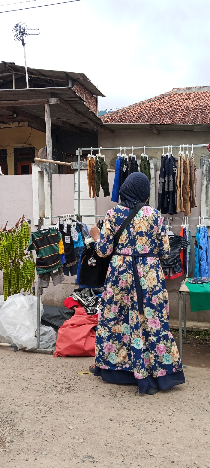 A woman wearing a floral dress and a hijab stands in front of a clothing rack at a market in Kecamatan Kutawaringin, Indonesia. The rack is filled with assorted children's clothes, hanging on metal hangers. A green plastic table sits to the right of the rack. There are various objects surrounding the table and clothing rack, including red, black, and grey bags. Green beans hang from a metal pole in the background. There is a building in the background, with a brown tile roof. The pavement is dusty, with a dirt-like surface. The woman is looking down at the clothing on the rack.