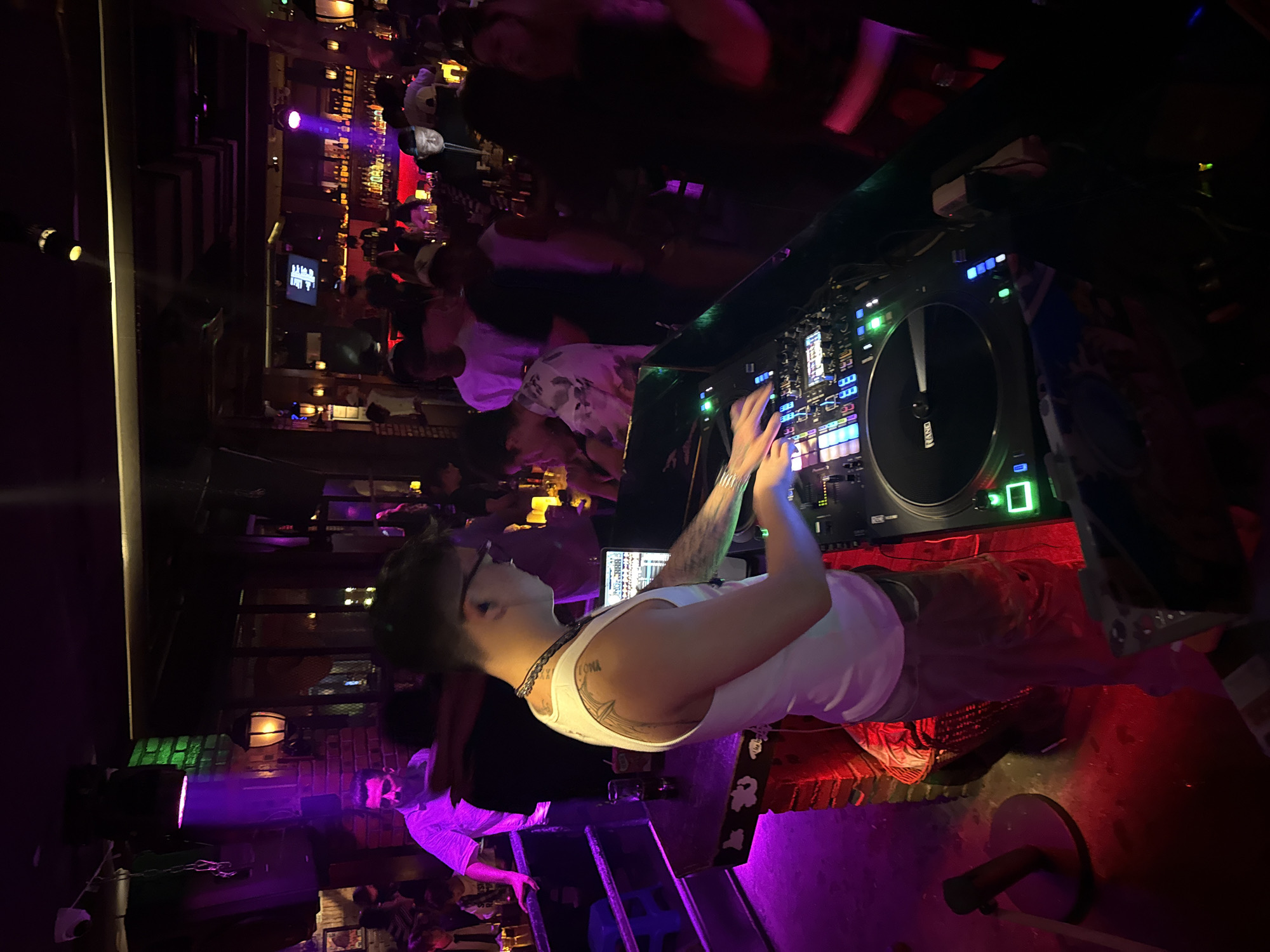 The image depicts a nightclub environment. A male DJ, wearing a white tank top and glasses, is seen from the side, actively mixing music on a DJ console which features a turntable and various electronic controls. He has tattoos on his left arm and shoulder. The club is dimly lit with vibrant purple and red lighting. In the background, a crowd of people are visible, some standing at a bar area lined with bottles. The overall atmosphere suggests a lively social gathering.
