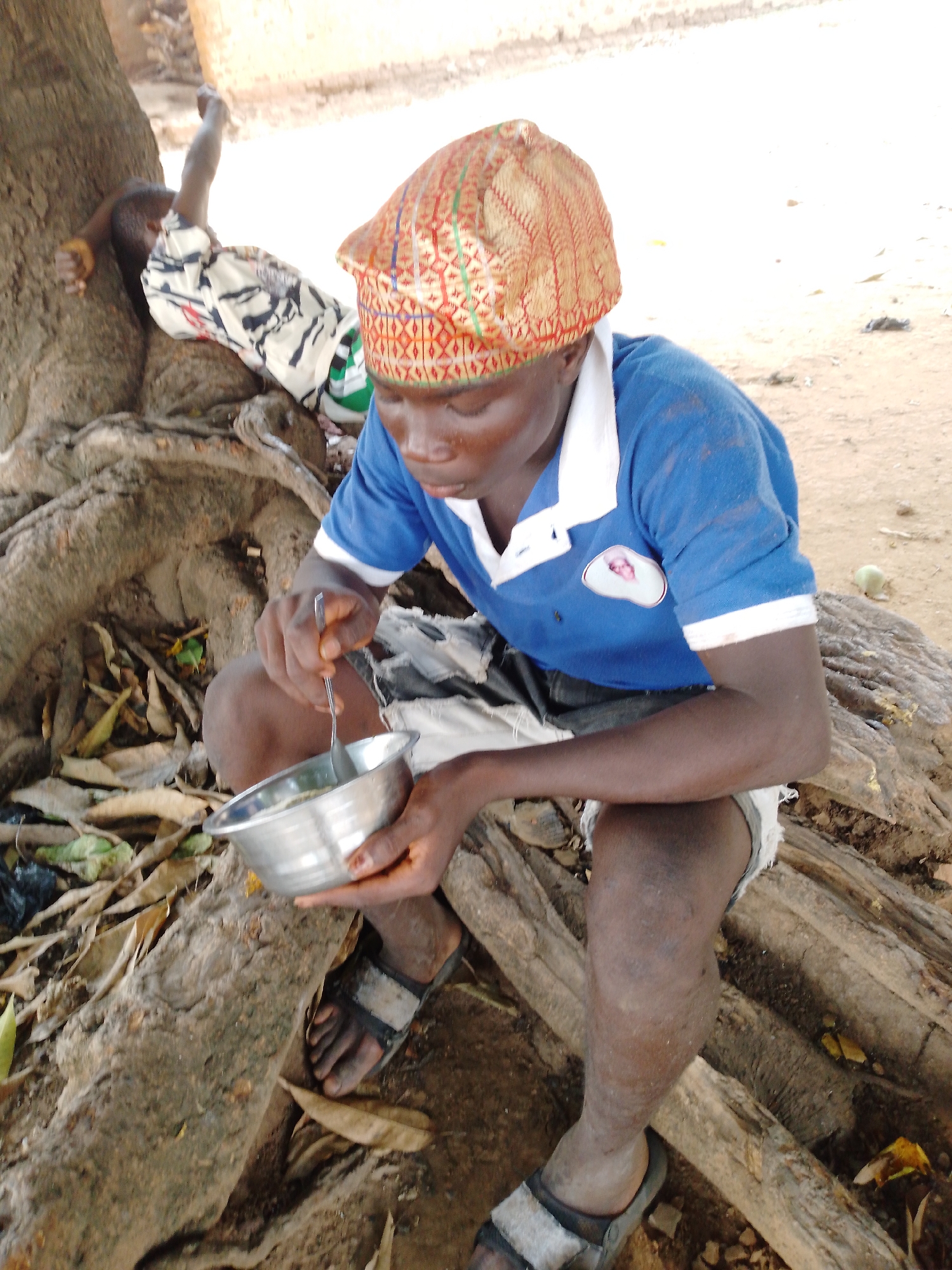 An outdoor, eye-level shot captures a young person eating while seated among the prominent roots of a large tree. The individual, appearing male, is positioned centrally, wearing a blue polo shirt with a white collar and trim, dark shorts that appear distressed, and dark sandals with light-colored straps. A distinctive patterned hat, possibly a Kufi or similar traditional cap, adorned with red, gold, and green designs, covers his head. He holds a silver-colored metallic bowl in his left hand and uses a spoon with his right hand to eat its contents, focusing intently on his meal. A small, blurry badge or logo, appearing as a white oval with a faint pinkish image, is visible on his blue shirt.
In the background to the left, another person, possibly a child, is partially visible, leaning or resting against the tree trunk, partly covered by a white and black patterned cloth. The setting features dusty, uneven ground covered with dried leaves and some scattered debris. Large, gnarled tree roots dominate the foreground and mid-ground, providing a natural seating area for the main subject. In the distant background, a light-colored building or wall is visible under natural daylight. The overall bright, soft lighting suggests it is daytime. The scene depicts a moment of daily life in an outdoor, likely rural or semi-urban, environment. No specific emotions are clearly discernible from the primary subject, who is engrossed in eating. No discernible text is visible beyond the unreadable logo. The location context is Akwanga, Nigeria.