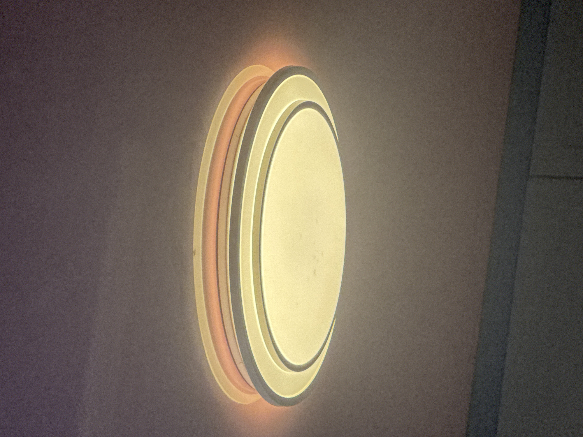 The image shows a close-up, low-angle shot of a modern, circular ceiling light fixture. The light is illuminated, casting a warm, yellow glow downwards and a softer, pinkish-orange hue around its edges onto the ceiling. The fixture appears to have multiple concentric rings, with the innermost one being the primary light source and the outer rings adding a decorative element and ambient lighting. The ceiling is textured and a dark, out-of-focus area is visible at the bottom of the frame, possibly indicating furniture or a lower part of the room. The overall impression is one of contemporary interior lighting. The location of Bohunove, Ukraine is not depicted or inferable from the provided image.