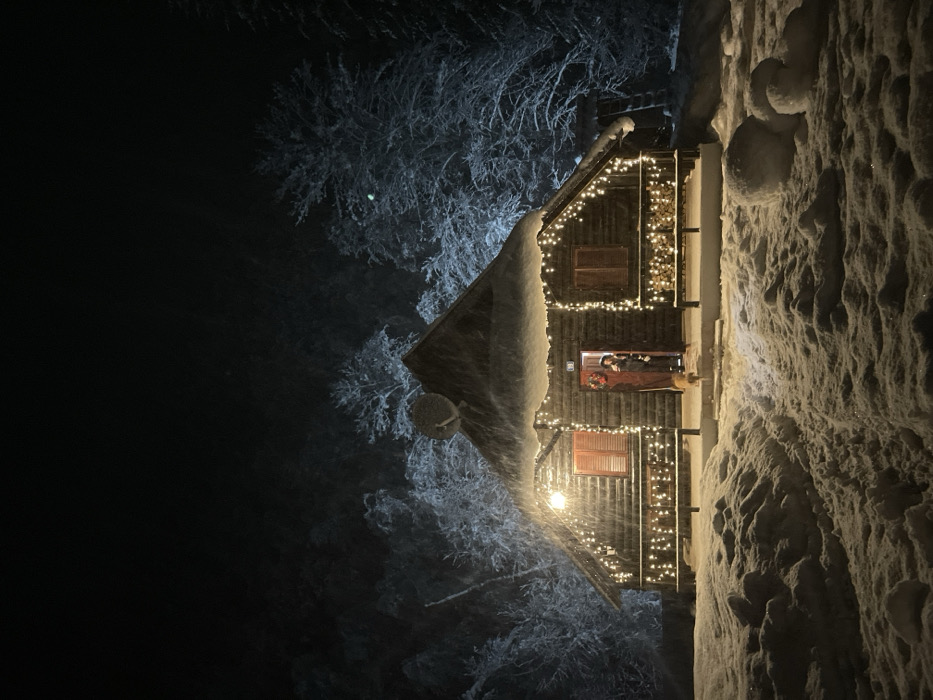 The image depicts a charming wooden cabin or lodge, festively adorned with warm string lights, set against a dark, snowy night in Lugovi, Montenegro. The original image is rotated 90 degrees clockwise; when viewed correctly, the cabin stands upright amidst a winter wonderland.
The cabin, constructed from dark timber, is blanketed in thick snow that covers its roof, balcony railings, and the surrounding ground. Numerous small, glowing yellow lights are strung along the cabin's exterior, illuminating the porch area and creating a cozy, inviting atmosphere. Two windows with closed wooden shutters are visible on the facade.
On the illuminated porch, a person is seen standing near an open doorway, wearing a dark coat and hat, suggesting cold weather. Their face is indistinct due to the distance and lighting. Adjacent to the person, a small, light-colored dog is also visible. A red wreath, likely festive, hangs near the entrance. Stacked logs of wood are piled on the porch, and a satellite dish is mounted on the cabin's left side.
The scene is dominated by a heavy snowfall, with snow-laden trees appearing ghostly white in the dark background. The ground is covered in deep, textured snow. The overall impression is one of a tranquil, cold winter evening, possibly during the holiday season, with the warm lights of the cabin providing a beacon of comfort. No visible text is present in the image.