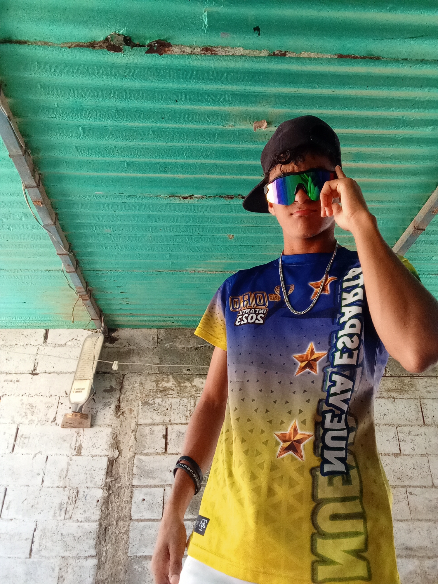 A young person wearing a blue and yellow shirt with star designs, white shorts, sunglasses, and a black cap is standing underneath a corrugated metal roof in a location within the city of Juan Griego, Venezuela. The shirt has "Oro Infantil" printed on it and a silver chain around the neck. The wall behind the person is made of white bricks. A light fixture is attached to the wall.