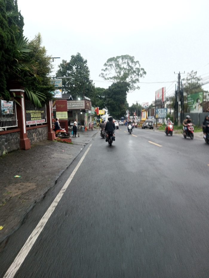 The image captures a street scene in the city of Chagzam_Pam, Bhutan, under an overcast sky. The foreground and midground feature a wet, dark asphalt road, indicating recent precipitation, with a solid white line delineating the left edge. Faint yellow dashed lines are visible further down the road. Numerous motorcycles and scooters, ridden by individuals wearing helmets, are traveling away from the viewer in the road's center and right lanes. A dark-colored car is also present in the distance.
To the left of the road, a raised concrete sidewalk is bordered by a low wall constructed from stone and dark red brick, topped with lush green foliage including conifers and palm fronds. A red and white scooter is parked on this sidewalk, with one individual seated on it. Other pedestrians stand near storefronts behind the sidewalk, including one person in a dark top and light trousers. Commercial buildings line the left side, displaying signs such as "Suryz," "SATE MAHANGGI BARUASH," "SELAMAT DATANG DI DATUK LIA PEULUS," and "ATM BANK..."
In the distance, on the right side of the road, a prominent sign for "SETIA HOTEL" is visible above additional commercial structures and utility poles with numerous overhead wires. Large trees are interspersed among the buildings in the background.