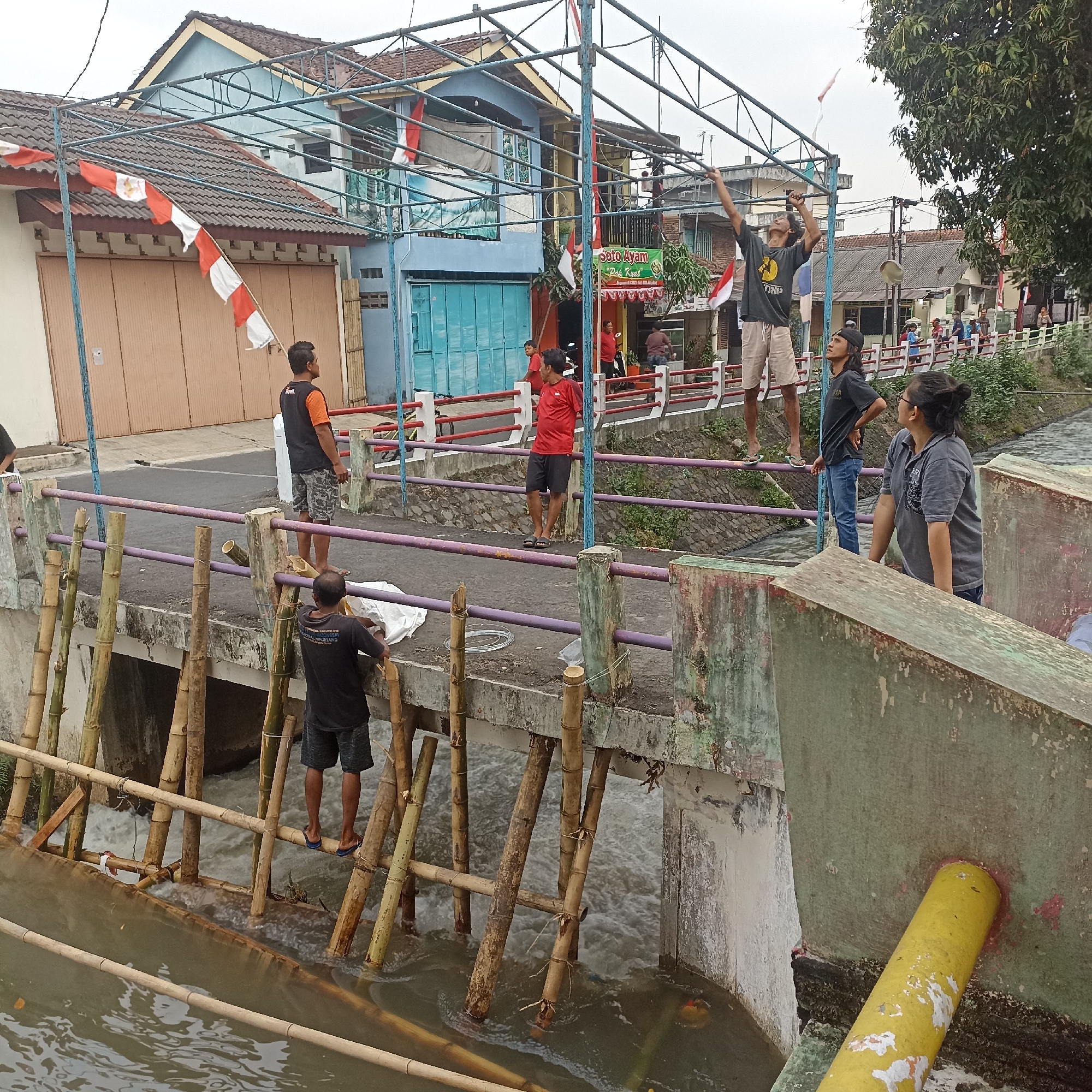 In Magelang, Indonesia, on a cloudy day, several individuals are gathered on a bridge spanning a river. The bridge railing is purple, with red and white decorative elements. A metal frame structure is erected over the bridge, adorned with red and white Indonesian flags. Several people are actively working on or near this structure.
On the bridge, a man in an orange and black vest stands looking to his right. To his left, another man in a red shirt and black shorts looks forward. A third man, in a black shirt and camouflage shorts, stands on the bridge's edge, looking up. Further left, a man in a black t-shirt and jeans stands with his hands on his hips. A woman in a grey polo shirt and glasses stands near the edge, looking to her right.
Below the bridge, bamboo poles have been erected in the river, likely as a temporary reinforcement or part of a construction. The river water is flowing briskly. On the bridge itself, two men are working on the bamboo structure. One is in a black shirt and shorts, and the other is in a black shirt with a white logo and dark shorts.
The background shows buildings with tiled roofs, some painted blue and light yellow, with cream-colored garage doors. A sign reading "Soto Ayam Pak Kyai" is visible on one of the buildings. Trees are present on the right side of the image. The overall scene suggests an ongoing event or preparation near the river in a semi-urban area of Magelang.