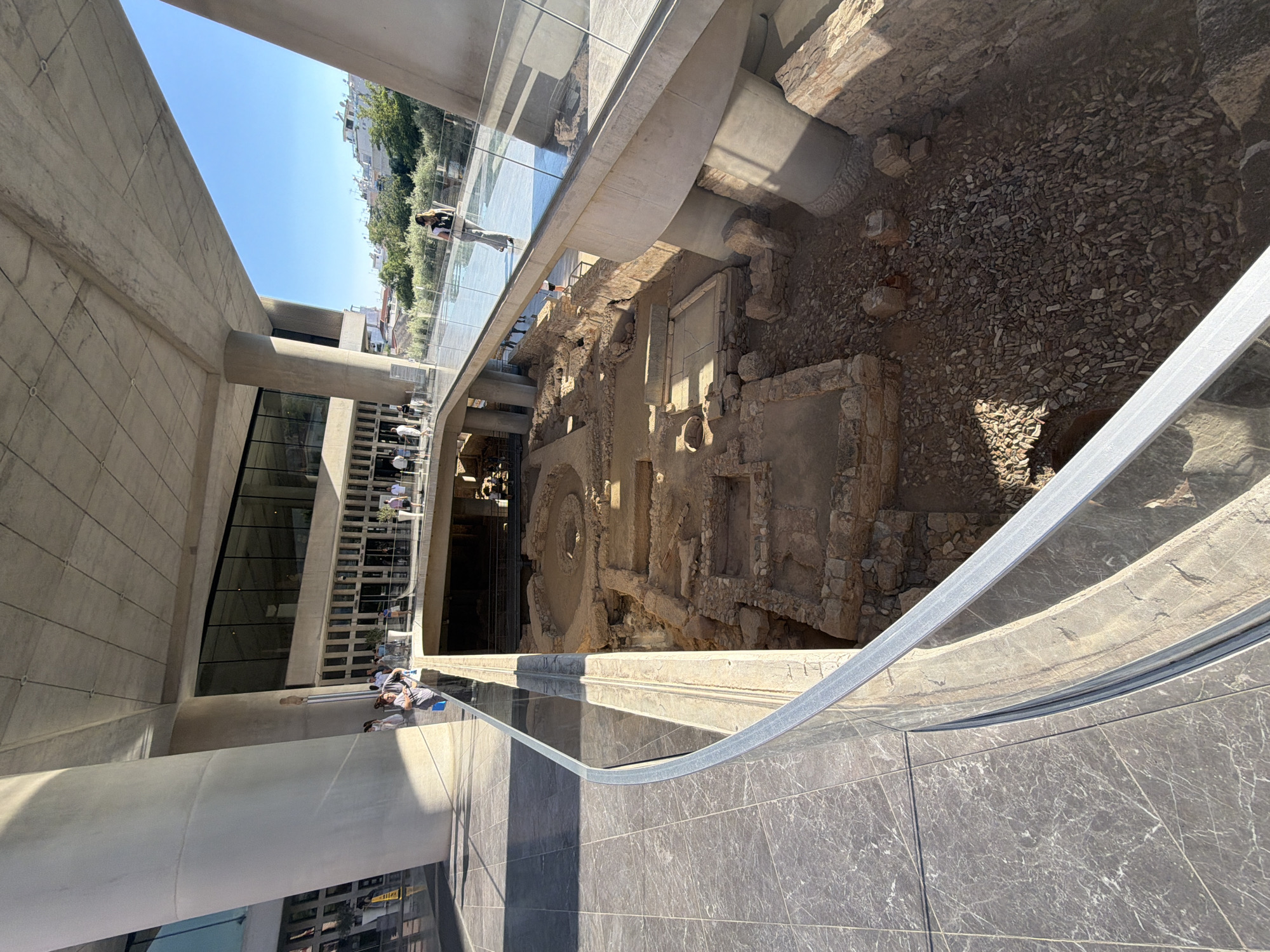 The image depicts an outdoor scene within a modern architectural structure that offers a view of an archaeological excavation site. Several people are visible, some standing and observing the ruins, while others appear to be walking along a glass-enclosed walkway. The walkway has a metal railing, reflecting the surrounding environment.
The archaeological site features ancient stone structures, including walls, foundations, and what appears to be a circular foundation or well. The ground within the excavation is covered with loose stones and dirt.
The setting suggests a museum or historical site with a contemporary design, possibly an extension or viewing platform built over or adjacent to the ruins. The bright sunlight and clear blue sky indicate a daytime scene, likely in pleasant weather. The architecture includes large concrete elements and glass facades.
The people's attire suggests casual wear, suitable for a warm day. Their posture and orientation suggest they are engaged in observing and exploring the site.
The location is identified as Athens, Greece, which aligns with the presence of ancient ruins and the architectural style. The contrast between the modern building and the ancient remnants is a prominent feature. There is no visible text in the image.