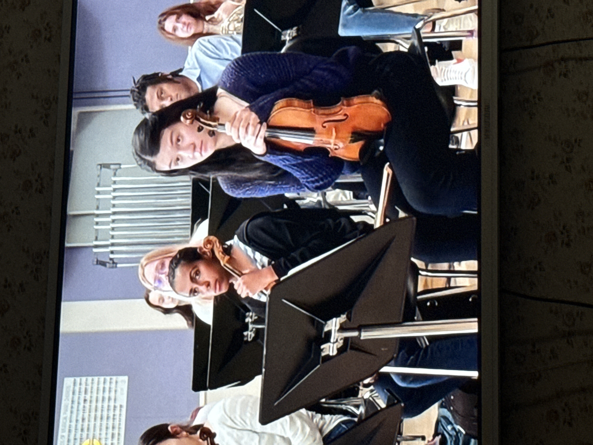 The image shows a group of people sitting in chairs, seemingly an orchestra or musical ensemble. Each person has a music stand and a violin. The people are looking forward. The background is a light purple color. Based on the context provided in the prompt, the location is likely a musical hall or a place where musical performances occur in Mytishchi, Russia. More information is needed to be certain of the precise location.