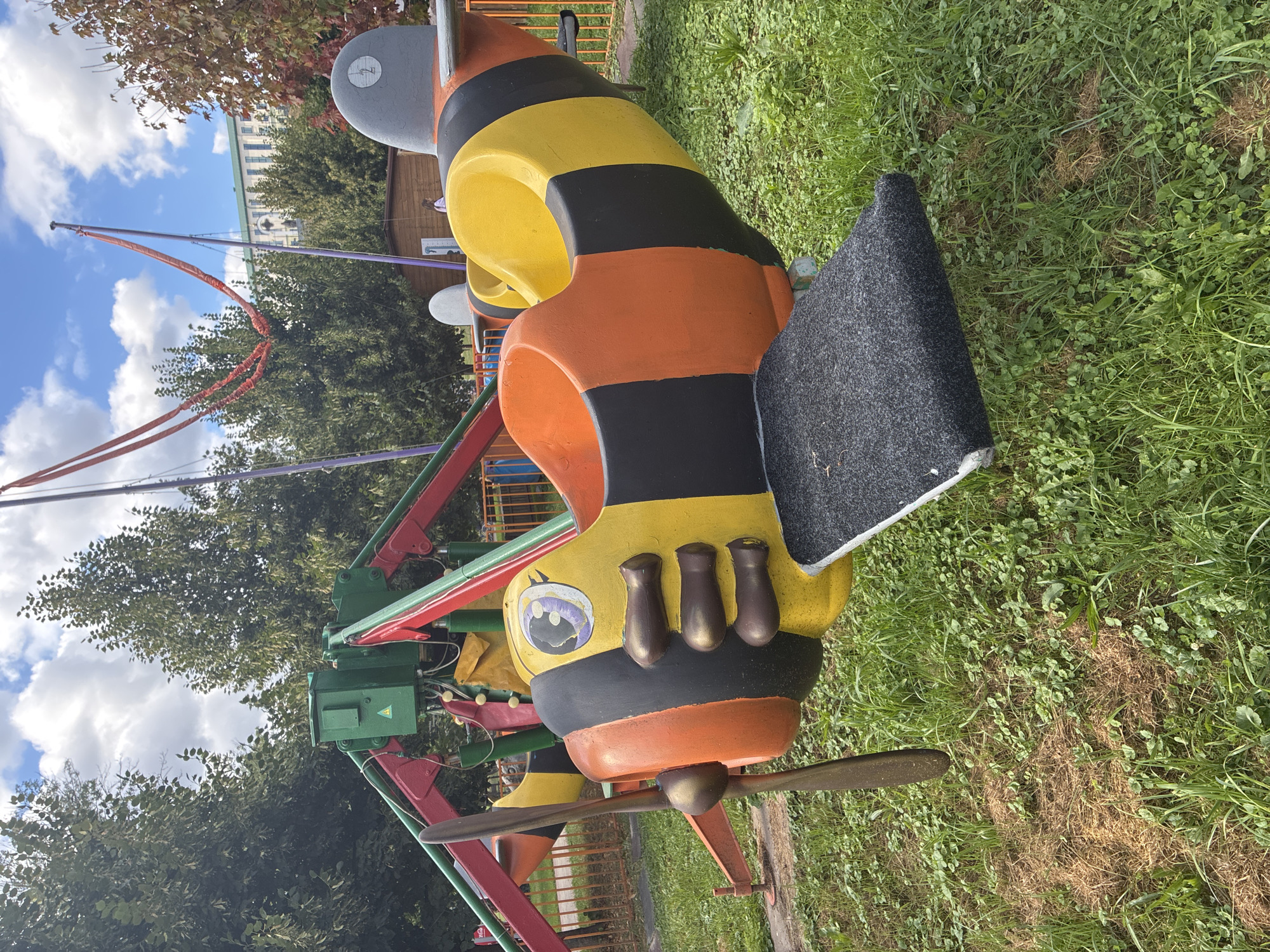 This is a medium-shot, eye-level photograph taken outdoors on a cloudy day in the city of Mahilyow, Belarus. The primary subject is a child's amusement ride shaped like a bumblebee airplane. The ride is yellow and orange with black stripes, and it features a propeller at the front. The seat is yellow and orange, with a black footrest. The ride is situated on a grassy area with some patches of dry grass. In the background, there are trees, part of another amusement ride with red and green metal structures, and a building. The sky is partly cloudy. There are no people clearly visible in the immediate foreground.