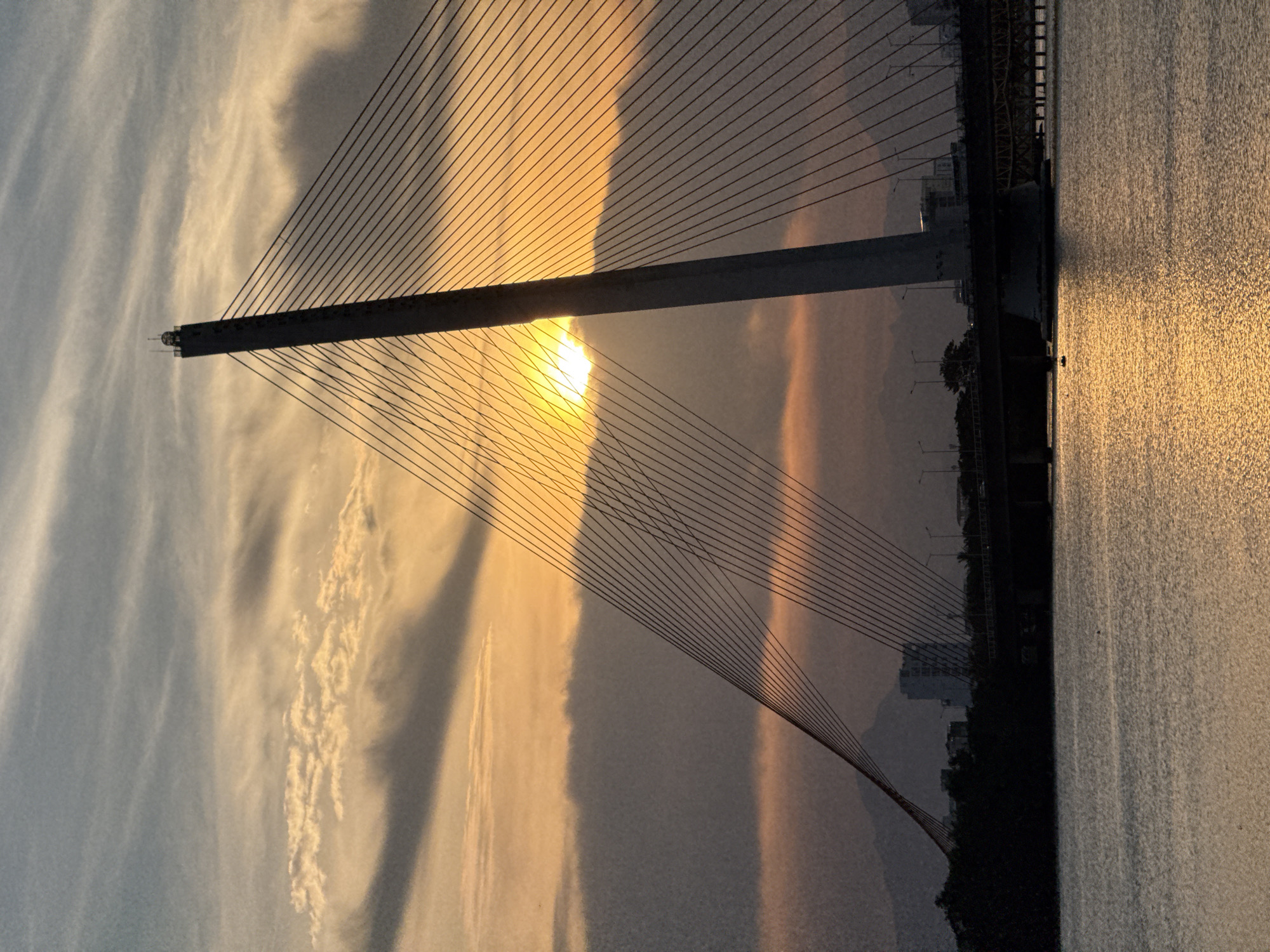 This is a photograph of the Dragon Bridge in Đà Nẵng, Vietnam, taken at sunset. The sun is low in the sky, casting a warm, golden light on the scene. The sky is filled with dramatic clouds, streaked with orange, yellow, and gray. The Dragon Bridge, a modern cable-stayed bridge, dominates the frame. Its tall, slender pylon and numerous cables are silhouetted against the colorful sky. The bridge's roadway is visible in the midground, with traffic lights and streetlights dotting the span. In the foreground, a body of water, likely the Han River, reflects the sunset's glow. The far bank of the river is lined with trees and buildings, with mountains visible in the distance. A small boat is present on the water, adding a touch of scale and human presence to the scene. The overall mood is peaceful and serene, with a sense of grandeur evoked by the impressive architecture and the stunning natural light.