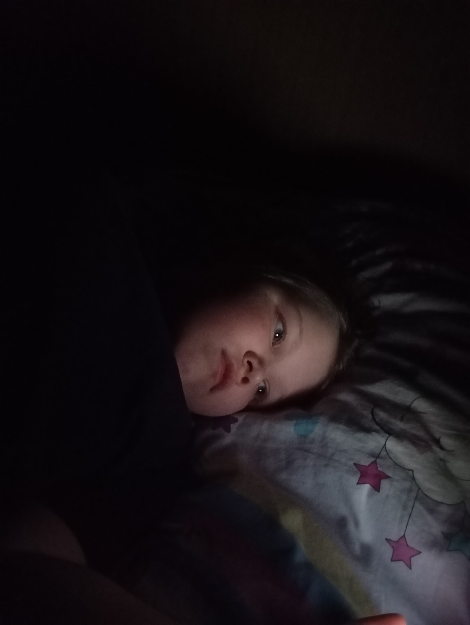 A child lies in bed on a patterned pillow and blanket, the light source on their face suggesting they are looking at a screen. The location is not discernible.
