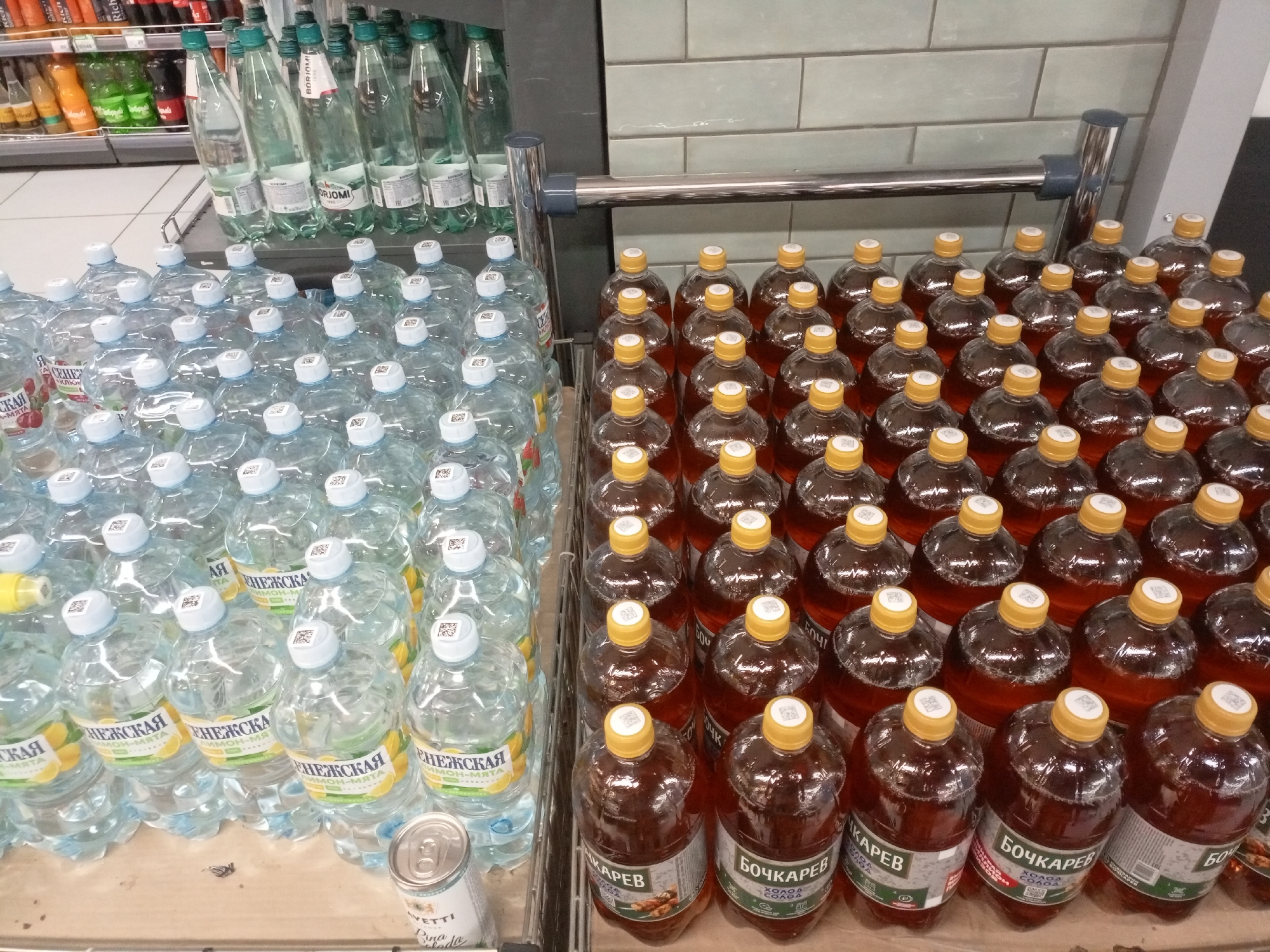 This image shows a retail display of bottled beverages in what appears to be a supermarket in Zheleznogorsk, Russia. The primary subjects are rows of plastic and glass bottles filled with various liquids, predominantly water and darker-colored beverages. On the left side of the display, a variety of clear plastic bottles with white or light-colored caps are arranged. Some of these bottles have labels in Russian that include Cyrillic text, such as "ЕНЕЖСКАЯ" and "ЛИМОН-МЯТА," indicating a lemon-mint flavored drink. Adjacent to these, a silver can with partial text is visible.
To the right of the clear bottles, there are rows of glass bottles containing a amber-colored liquid. These bottles have yellow caps and labels with Russian text, including "БОЧКАРЕВ" and "ХОЛОД. НАП." The background shows shelves stocked with more beverages, including bottles with green caps and labels like "BORJOMI." The setting is indoors, with a tiled wall and a metal retail shelving unit visible behind the bottles. The lighting suggests it is daytime, likely during business hours. No people are visible in the image, focusing the attention solely on the products. The overall scene depicts a well-stocked beverage section of a store.