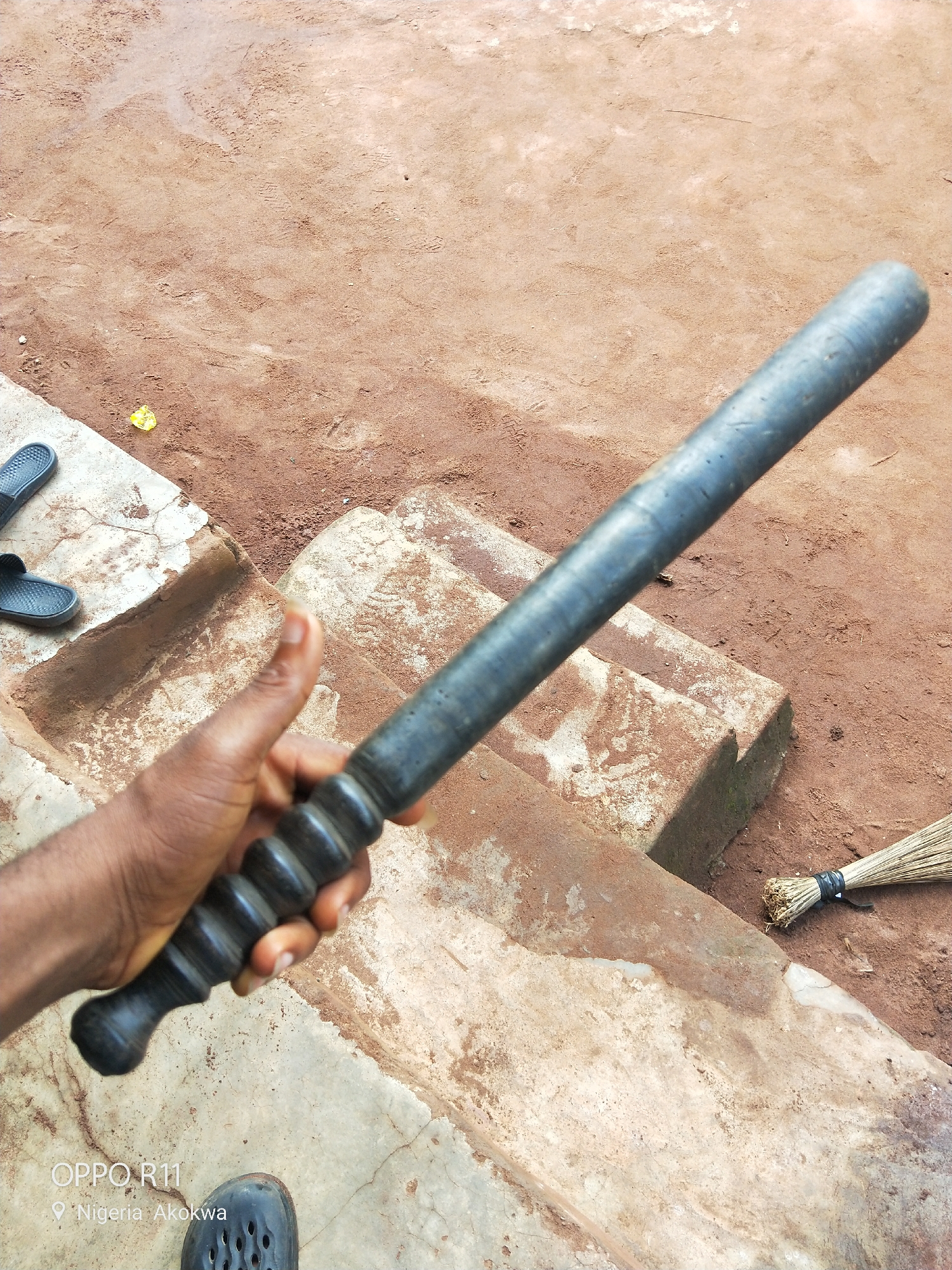A person's hand holds a dark-colored baton with a ribbed handle. The baton is positioned diagonally across the frame, with the handle closest to the viewer and the tip extending upwards and to the right. The hand appears to be dark-skinned, and the fingernails are visible.
The background consists of a dirt ground with some texture, possibly indicating dried mud or footprints. To the left, there are concrete steps or ledges, with a pair of dark-colored flip-flops placed on the uppermost surface. Further to the right, a broom made of natural fibers is leaning against the edge of the concrete structure, its bristles fanned out on the dirt ground.
The scene appears to be outdoors, with natural lighting. The overall impression is one of a casual or possibly utilitarian setting. The colors are predominantly earthy tones of brown and gray, with the dark baton and flip-flops providing contrast.
Visible text at the bottom left of the image reads "OPPO R11" and "Nigeria Akokwa," indicating the phone model used for the photograph and the geographical location. There are no explicit signs of activity or event taking place, beyond the implied act of holding the baton. The time of day and weather conditions are not discernible.
