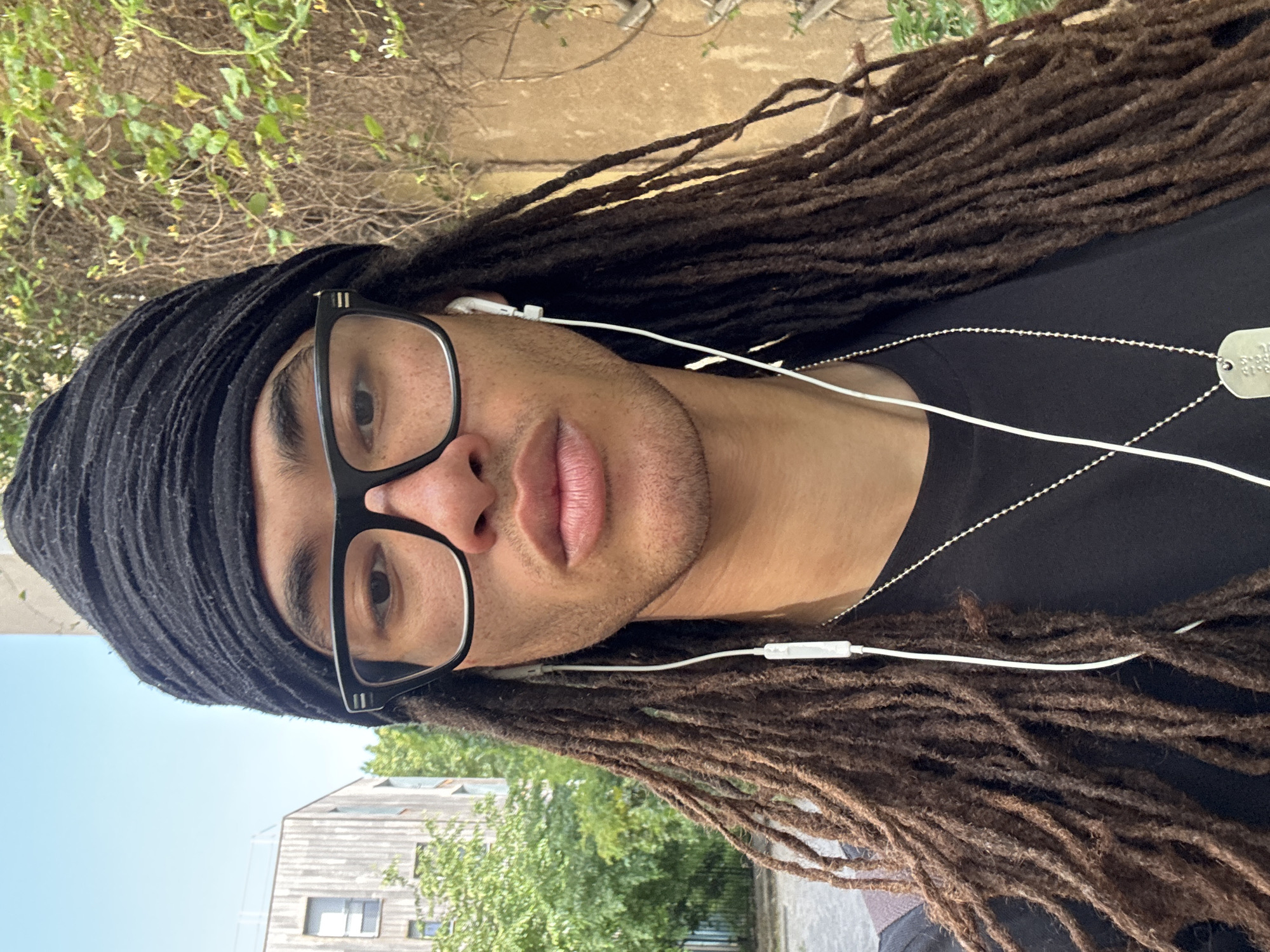 A portrait-style photograph shows a light-skinned person with brown dreadlocks. They wear a black head wrap and black-framed glasses. White earbuds are visible. A black t-shirt is worn with a silver chain and dog tags. The background includes foliage, a building, and a blue sky, suggesting an outdoor setting. The location cannot be determined.