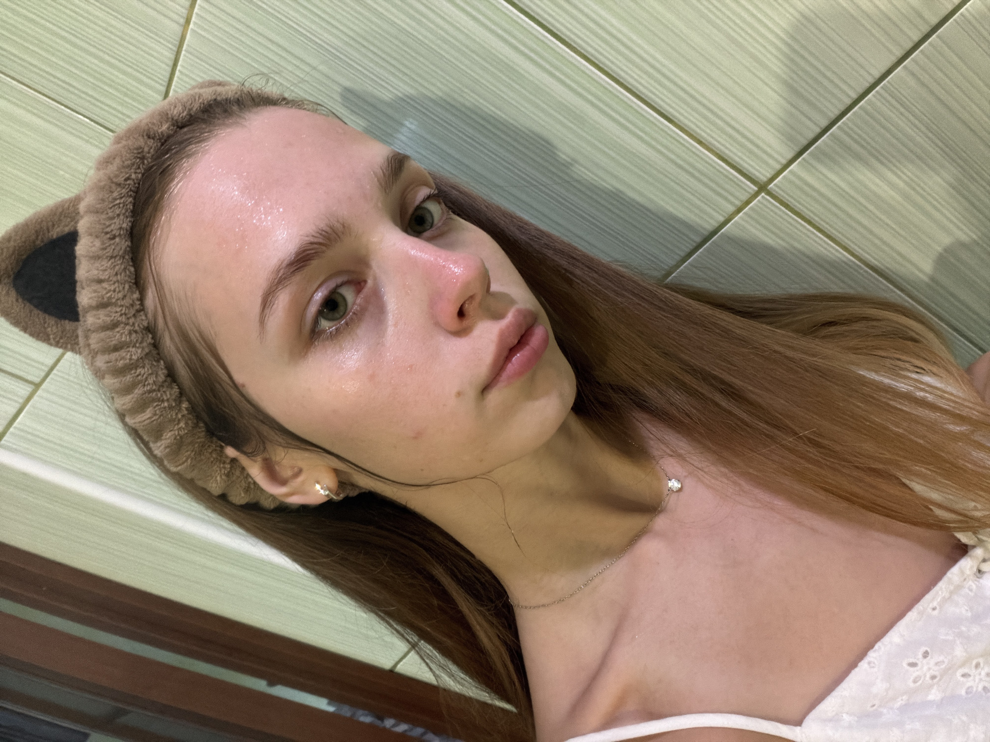 A young woman with light skin and light brown hair is shown in a close-up selfie. She is wearing a fuzzy brown headband with cat ears. Her eyes are green and she has a clear complexion, with some shine on her forehead and cheeks. She is wearing a thin silver necklace with a small diamond pendant. She also has a small diamond stud earring in her visible ear. The background appears to be a wall covered in light green, striped tiles. The lighting suggests it might be daytime, and the overall mood is casual and relaxed. She is not actively engaged in any particular activity, but rather posing for a photograph. There is no visible text in the image.