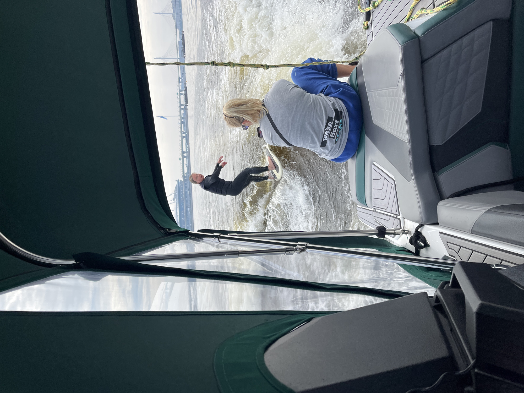 A sunny day over the Gulf of Finland, with a modern wake boat cutting through the choppy water. Two women are onboard, one actively wake surfing, the other sitting and watching. The surfer, wearing a black wetsuit and a life vest, is crouched low on her board, riding the wake with practiced ease. The spectator, in a grey t-shirt and blue pants, is facing away from the camera, her blonde hair pulled back in a ponytail. She's holding a phone and appears to be recording the action. The boat's interior is visible, with plush grey and green seating. In the background, the majestic cable-stayed bridges of Saint Petersburg, likely the Zolotoy Bridge and the Western High-Speed Diameter, arch gracefully against the cloudy sky. The scene captures a moment of exhilarating recreation amidst the urban grandeur of Russia's northern capital.