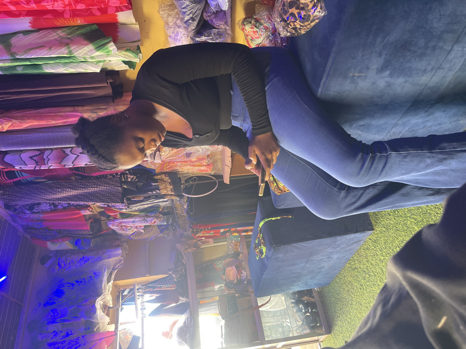 This image captures an interior scene within a retail clothing store or boutique in Samaru, Nigeria. A young adult female with dark skin and dark braided hair is prominently seated on a blue velvet ottoman, wearing a black long-sleeved top and blue flared jeans. She is focused on a smartphone held horizontally in both hands. The store's interior features walls lined with numerous garments of various colors and patterns, displayed on hangers and racks. Some merchandise, including what appears to be bags or accessories, is packaged in clear plastic and stacked. Shelving units on the left also hold various items. The floor is covered with green artificial turf. Overhead, blue LED strip lights illuminate parts of the space, while bright white light enters from an opening on the left side. An additional blue velvet ottoman is visible to the female's left, with a green and yellow beaded accessory resting on it.
