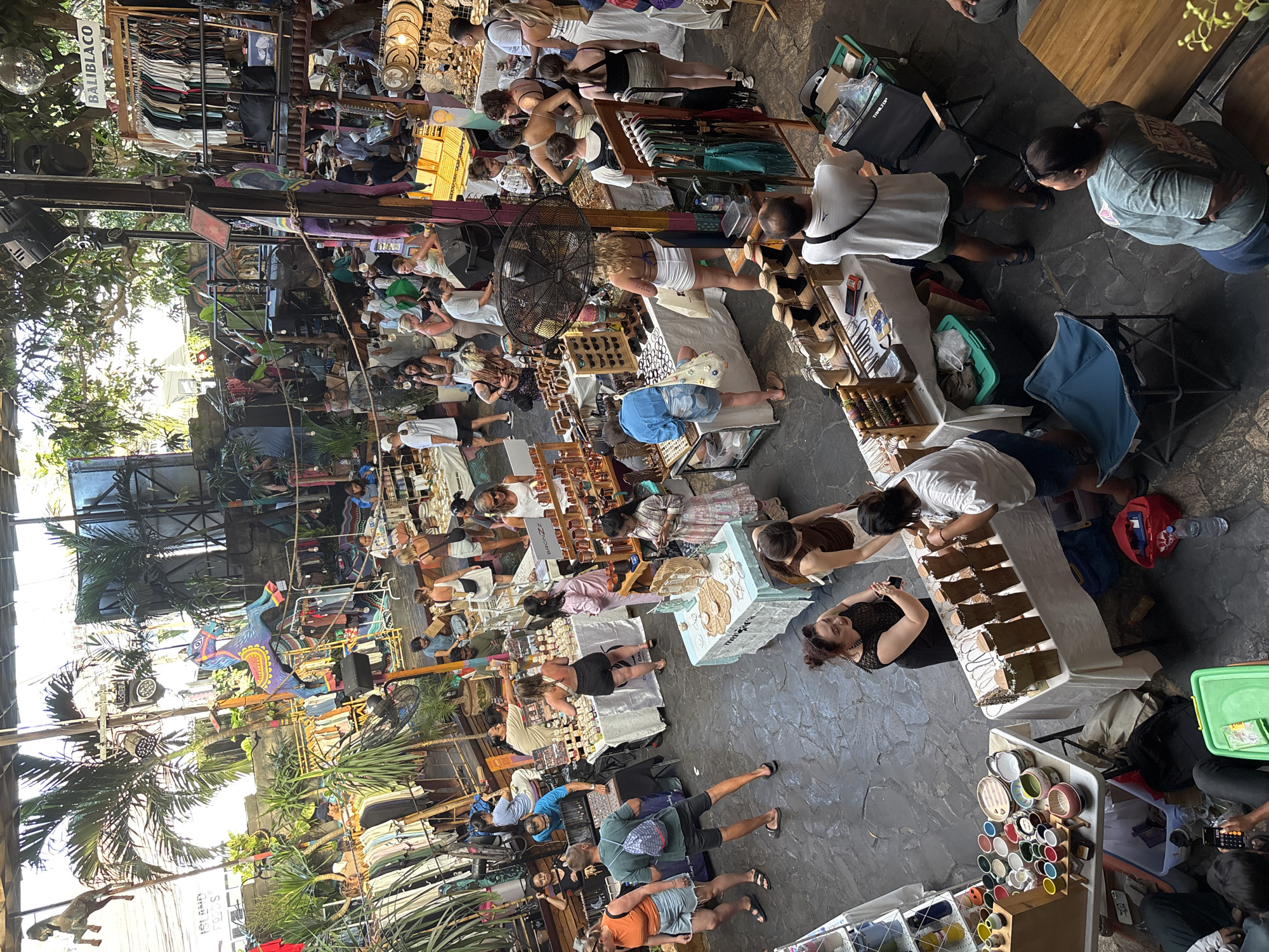 This image captures a lively scene at a market in Kuta Selatan, Indonesia, with a diverse crowd browsing various stalls. In the foreground, several individuals are engaged in shopping or interacting at tables laden with goods. To the left, a display of colorful ceramic cups and bowls is arranged on a white tablecloth-covered table. Adjacent to this, a man wearing a white t-shirt and dark shorts stands at a stall featuring necklaces and other jewelry. Further right, a woman in a dark top examines items at a stall with a white tablecloth, while another woman in a white top and a patterned skirt browses jewelry.
The mid-ground reveals more stalls, some with clothing racks showcasing various garments. A prominent feature is a large, colorful wooden horse decoration suspended above some of the stalls. Palm trees are visible throughout the market, suggesting an outdoor or semi-outdoor setting. A fan is positioned in front of a stall where a woman in a white top and black shorts is looking at items. In the background, more people are seen, some at higher levels, and various structures and decorations contribute to the bustling atmosphere. The overall impression is one of a vibrant marketplace filled with shoppers and vendors.