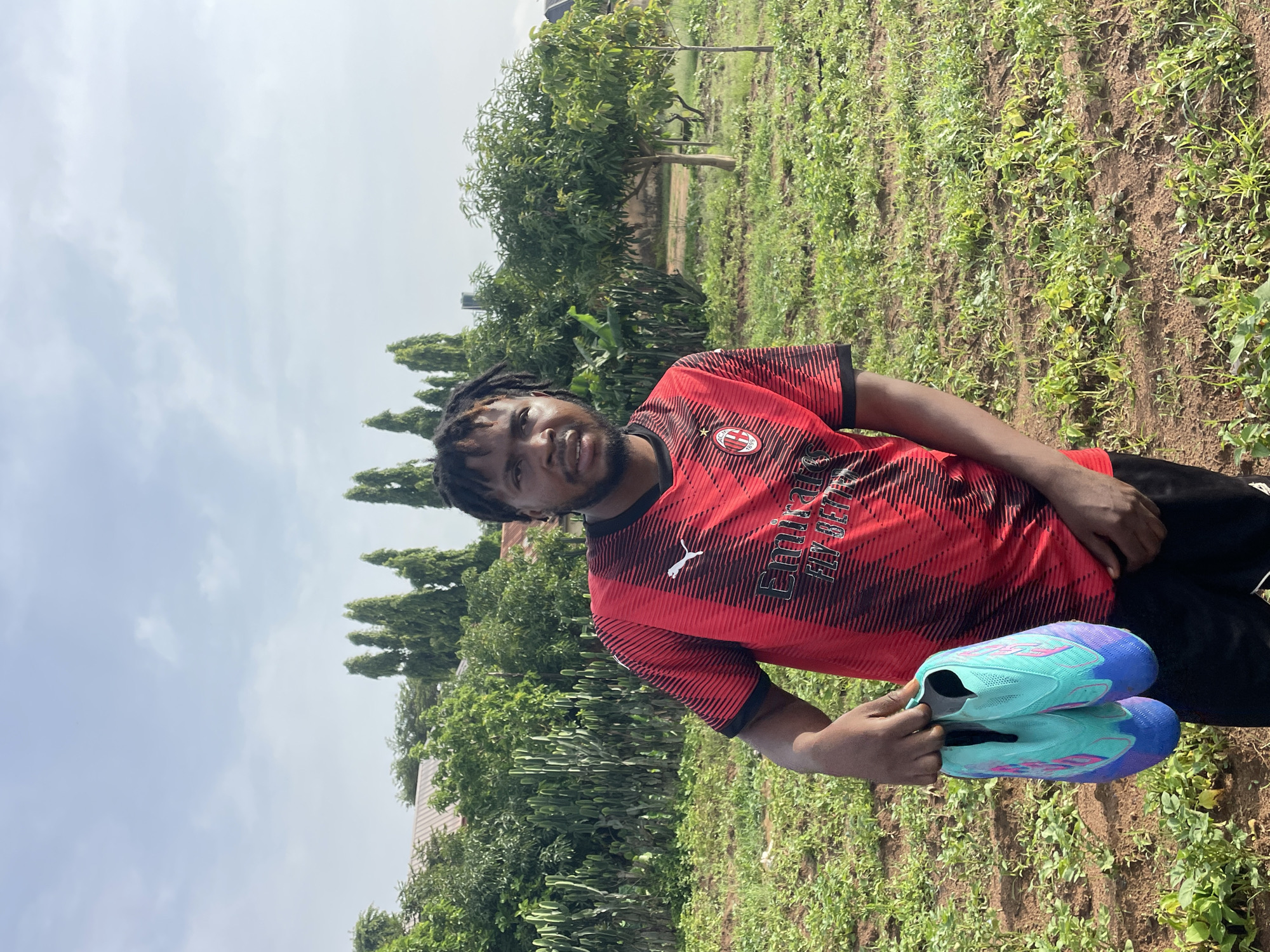 A man with dreadlocks, wearing a red and black AC Milan jersey and black shorts, stands in a field holding a pair of turquoise and purple soccer cleats. He is looking towards the camera. Behind him, a line of tall, slender trees stands against a cloudy sky. The field is covered in low-lying green plants and reddish-brown soil. In the background, there are houses and more trees. The image appears to have been taken outdoors during the daytime.