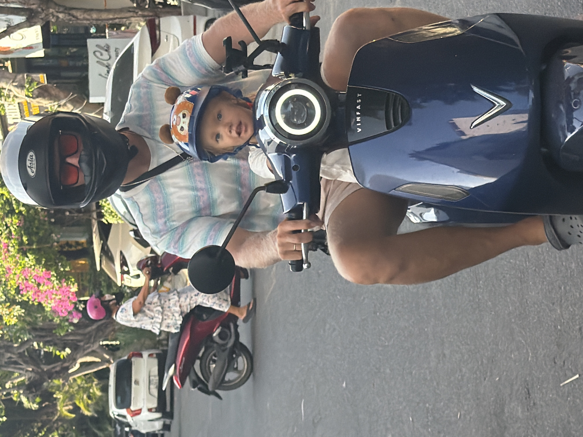 A man wearing a black helmet with red sunglasses and a striped t-shirt is riding a blue VinFast electric scooter. A young child wearing a blue helmet with a cartoon bear design is seated in front of him. The child is looking towards the camera with an open mouth. In the background, a woman in a white floral outfit and a pink helmet is riding a red scooter. White cars are parked along the street, and trees with pink flowers are visible. The scene appears to be a street in Nam Nha Trang Ward, Vietnam, under natural daylight.
