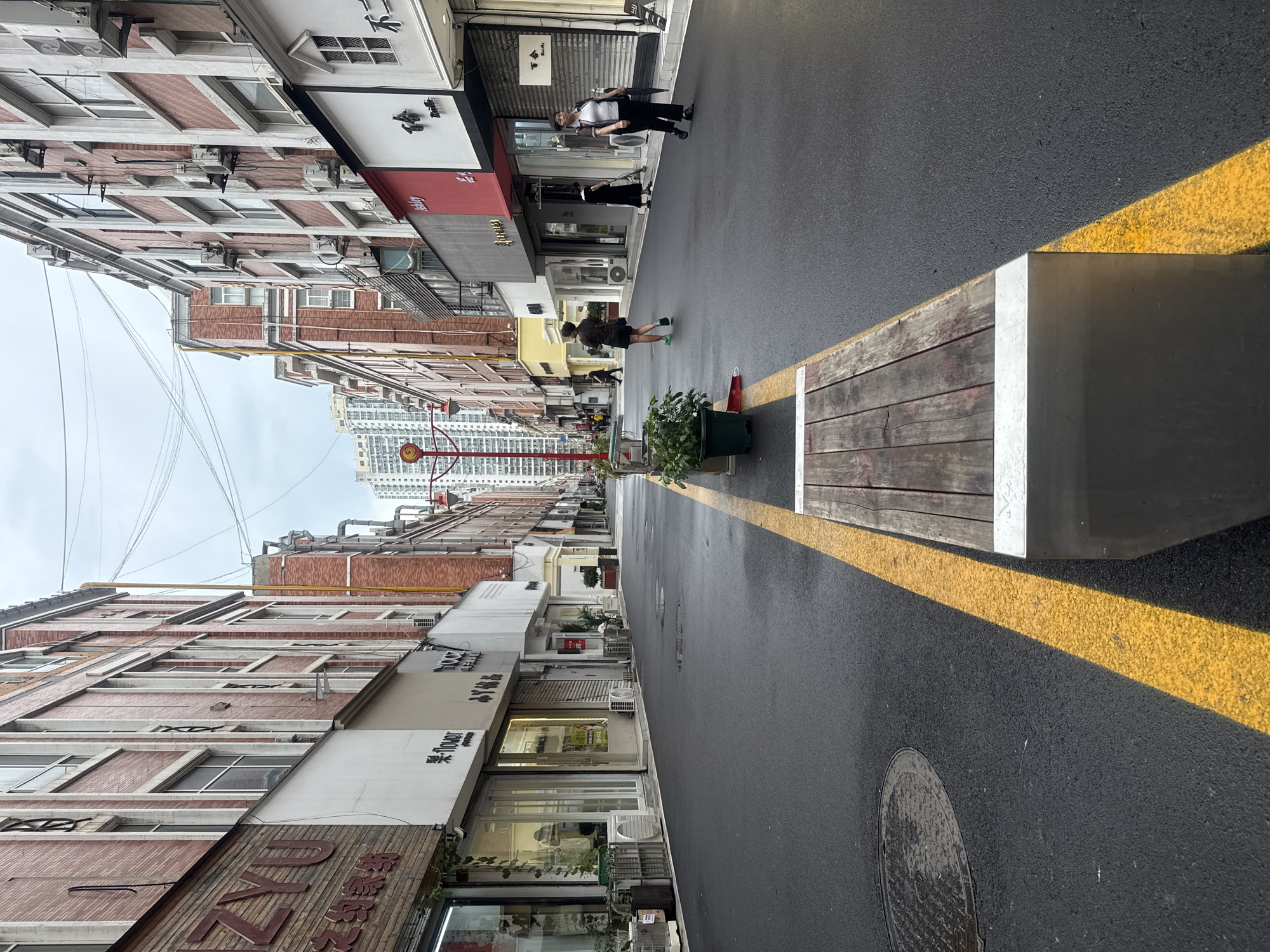 The image depicts a street scene in Qinhuangdao, China. On the left side of the street, brick buildings with white trim and numerous windows line the thoroughfare. Several shops with glass storefronts are visible, displaying various goods. A red sign with Chinese characters, partially obscured by a wooden facade, is prominent on one of the buildings. On the right side, similar brick buildings with white accents and windows also dominate the view, with shops and storefronts interspersed. The street itself is paved with asphalt and features a distinct yellow line running down its center, dividing the traffic lanes. A wooden bench with metal legs is positioned along the edge of the street, with a planter box containing a green plant next to it. Several individuals are present on the street. In the foreground, on the right, a woman with short dark hair, wearing a white shirt, black vest, and black pants, walks with an umbrella. Further down the street, another woman in a black dress is visible. On the left side of the street, a person with dark hair and a brown shirt is walking away from the camera. In the background, taller buildings, some with visible air conditioning units, rise into the sky, and a cloudy sky is overhead. The overall atmosphere appears to be that of a commercial street.
