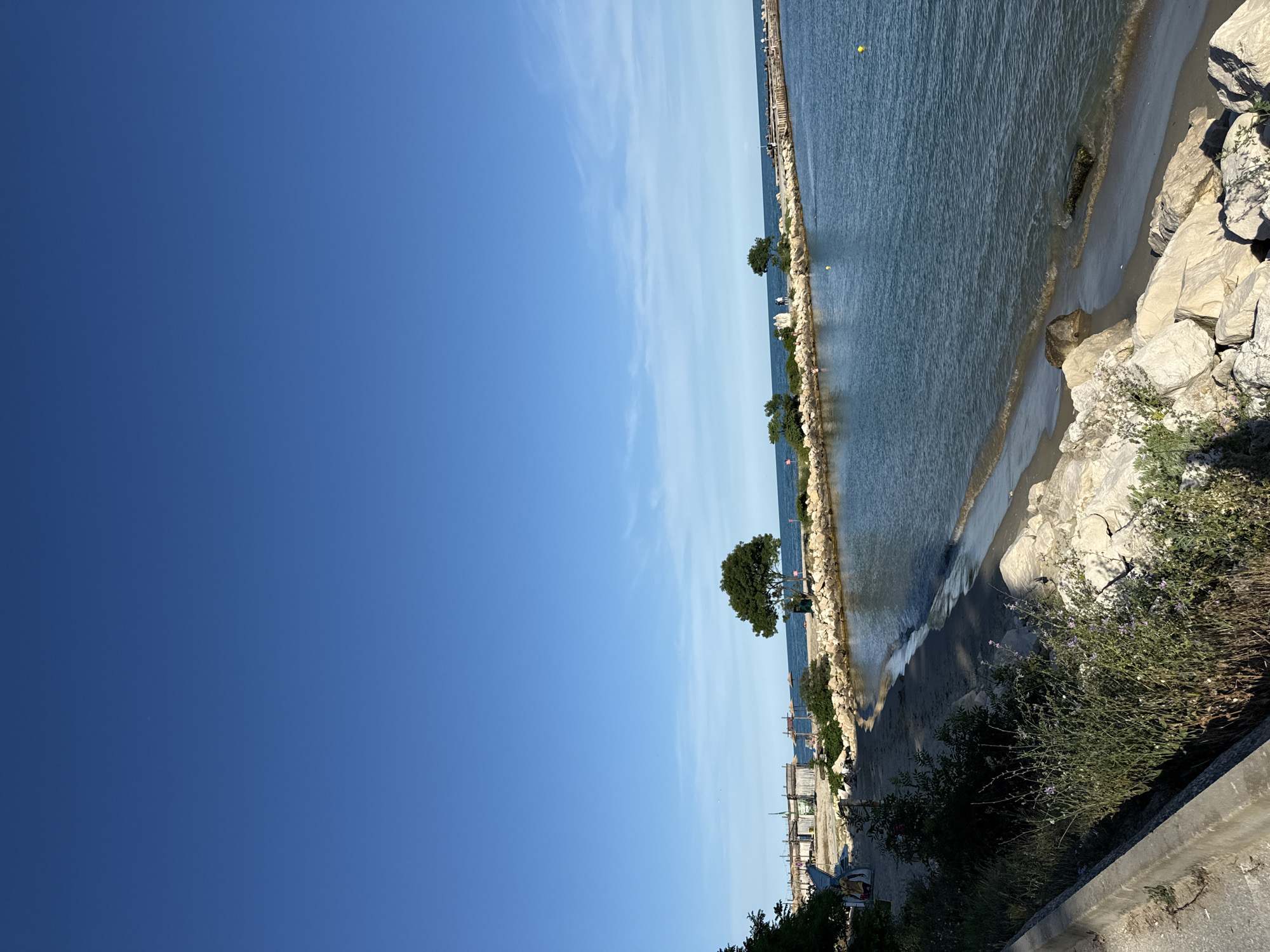 The image displays a coastal scene on a clear, sunny day. The foreground features a textured, concrete surface, suggesting a walkway or embankment. To the right, dense green bushes with small purple flowers grow, interspersed with rocks. A sandy beach stretches along the edge of the water, with gentle waves lapping at the shore. Large, light-colored rocks are scattered along the coastline.
Further out, the calm, blue water is dotted with a few small yellow buoys. A stone breakwater or jetty extends into the sea, lined with scattered greenery and a few trees. Small figures, presumably people, can be seen on or near the breakwater. In the distance, a pier or boardwalk stretches across the water, with some small structures visible on it. Along the shore, a line of simple, wooden beach huts or cabanas is present, with some beach chairs and umbrellas set up. A lone, mature tree stands prominently on the breakwater.
The sky is a vibrant, cloudless blue, indicating fair weather and likely daytime, possibly late morning or afternoon due to the clear light. The overall mood is peaceful and idyllic, evoking a sense of a relaxed day at the seaside. No visible text is present in the image.