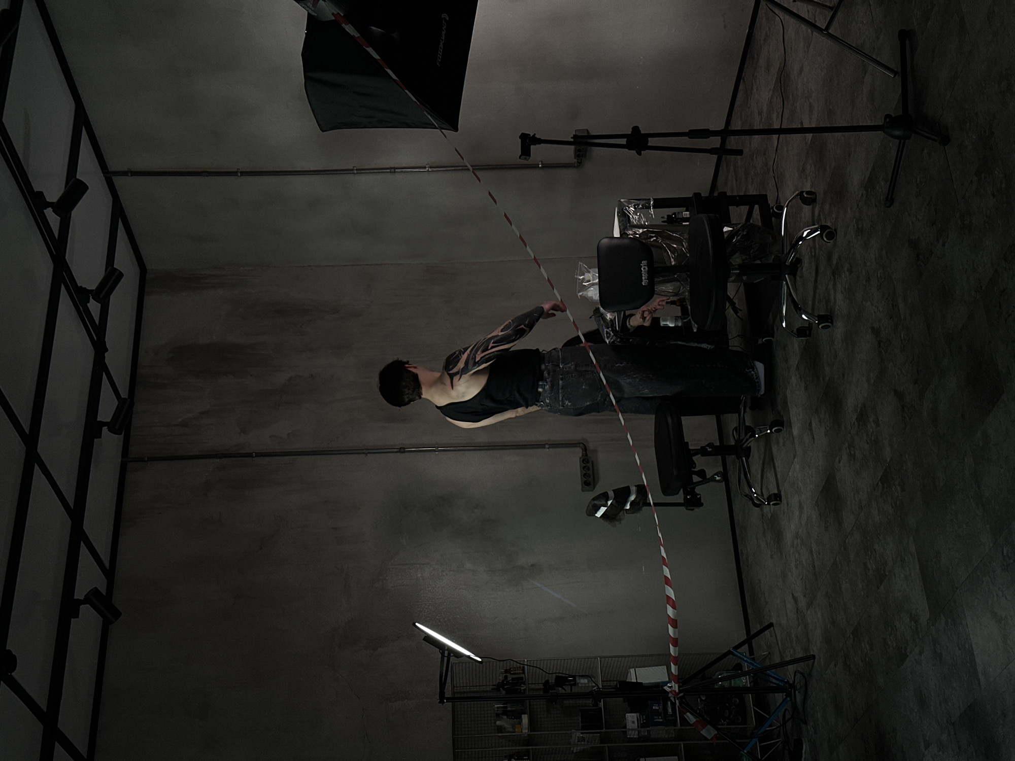 An interior shot, rotated 90 degrees counter-clockwise, depicts a person in what appears to be a tattoo studio or an industrial-style creative space in Moscow, Russia. The room features exposed concrete walls and floor, with visible metal conduit piping running along the upper sections of the walls.
In the mid-left of the frame, a person with dark hair, wearing a black tank top and dark trousers, stands, looking towards the right. Their left arm is extensively covered in black ink tattoos with intricate designs. To the right of the person, a professional setup includes a black office chair with "opsola" written on its headrest, and a table holding what appears to be tattoo equipment, some covered in plastic wrap.
Above the workstation area, a black softbox, possibly branded "FOTOKJIAN," hangs from the ceiling, indicating the presence of photographic or video lighting. Another bright, white LED light bar on a stand provides additional illumination from the lower-right of the frame. A red and white striped barrier tape runs diagonally across the image, seemingly segmenting the space.
Other notable details include a black metal grid structure with several spotlights on the left wall, a power strip with outlets visible on the right wall, and a metal shelving unit with various items stored on it in the lower-right. The scene suggests either a tattoo session underway, preparation for one, or a professional photo/video shoot related to tattooing or body art. The ambiance is utilitarian and focused, relying entirely on artificial lighting.