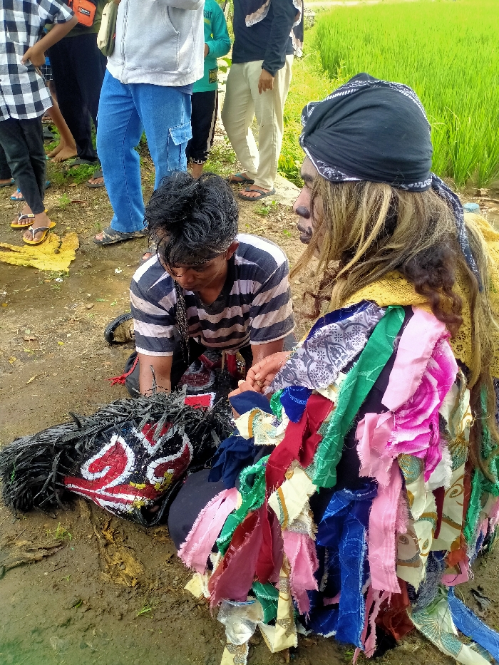 A young man wearing a striped shirt is kneeling on the ground in front of an older woman with long blonde hair, wearing a black headscarf and a long, colorful robe made of many scraps of fabric. The man is holding a red and black mask with a hairy face in front of the woman. There are several other people in the background. The scene is outdoors on a dirt path, with a field of tall green grass in the background. The location is near the coordinates latitude -7.52 and longitude 111.027, which is in Kecamatan Kedawung, Indonesia.