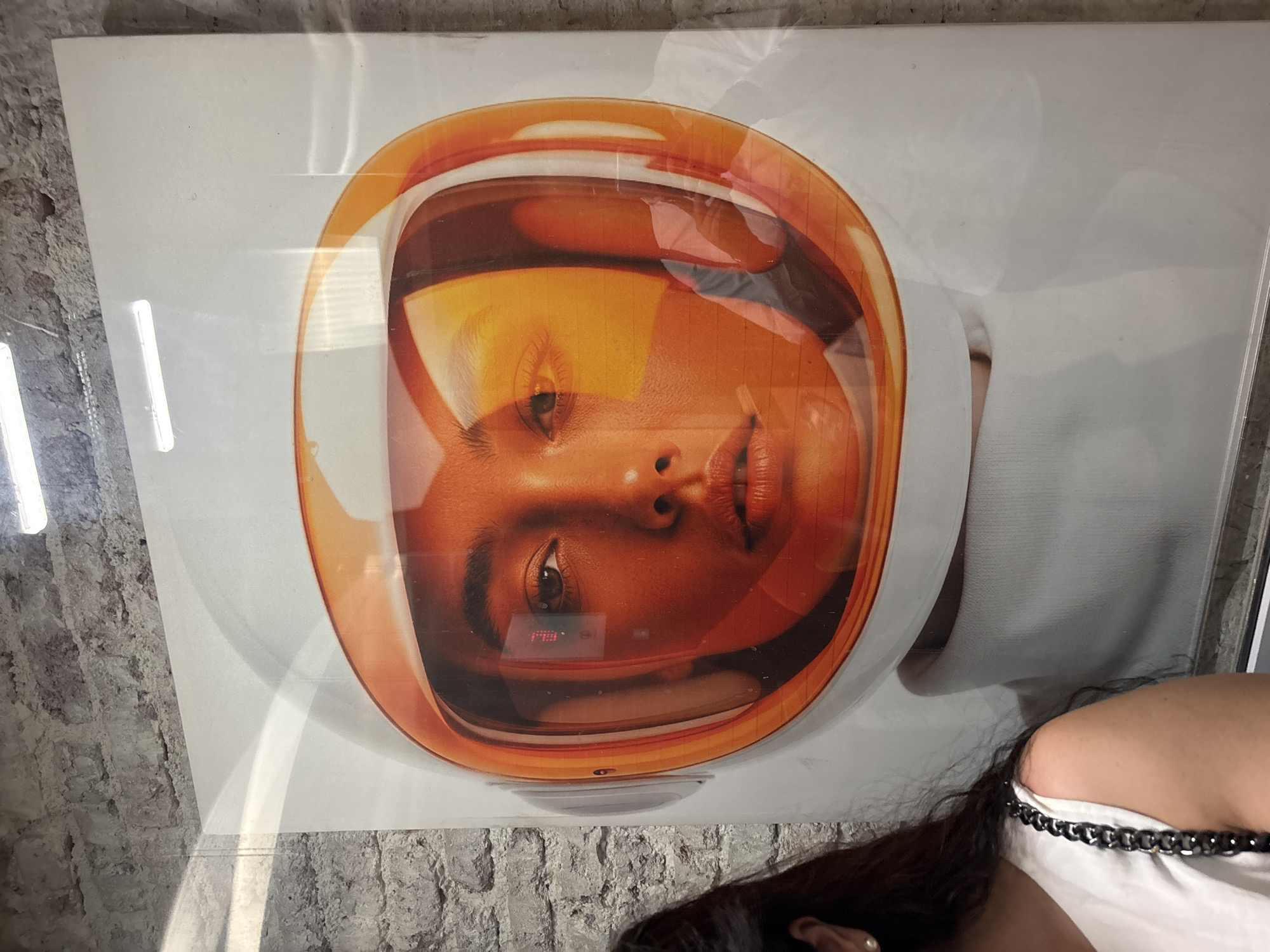 The image depicts a poster of a woman's face inside an orange-trimmed astronaut helmet. The poster is positioned vertically against a brick wall, likely indoors. A partial view of a person with long brown hair and a white top with a black chain strap is visible in the lower right corner of the frame. The location is in Mexico City, Mexico.