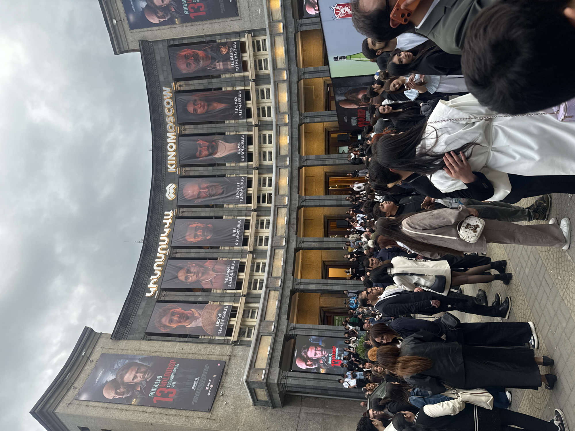 The image shows a crowd of people lined up in front of the Kinomoskva cinema in Yerevan, Armenia. Banners with Armenian inscriptions and headshot portraits of actors are hung along the facade of the building, and movie posters are placed on the sides. People of various ages and styles of dress are waiting outside the cinema. The sky is overcast.