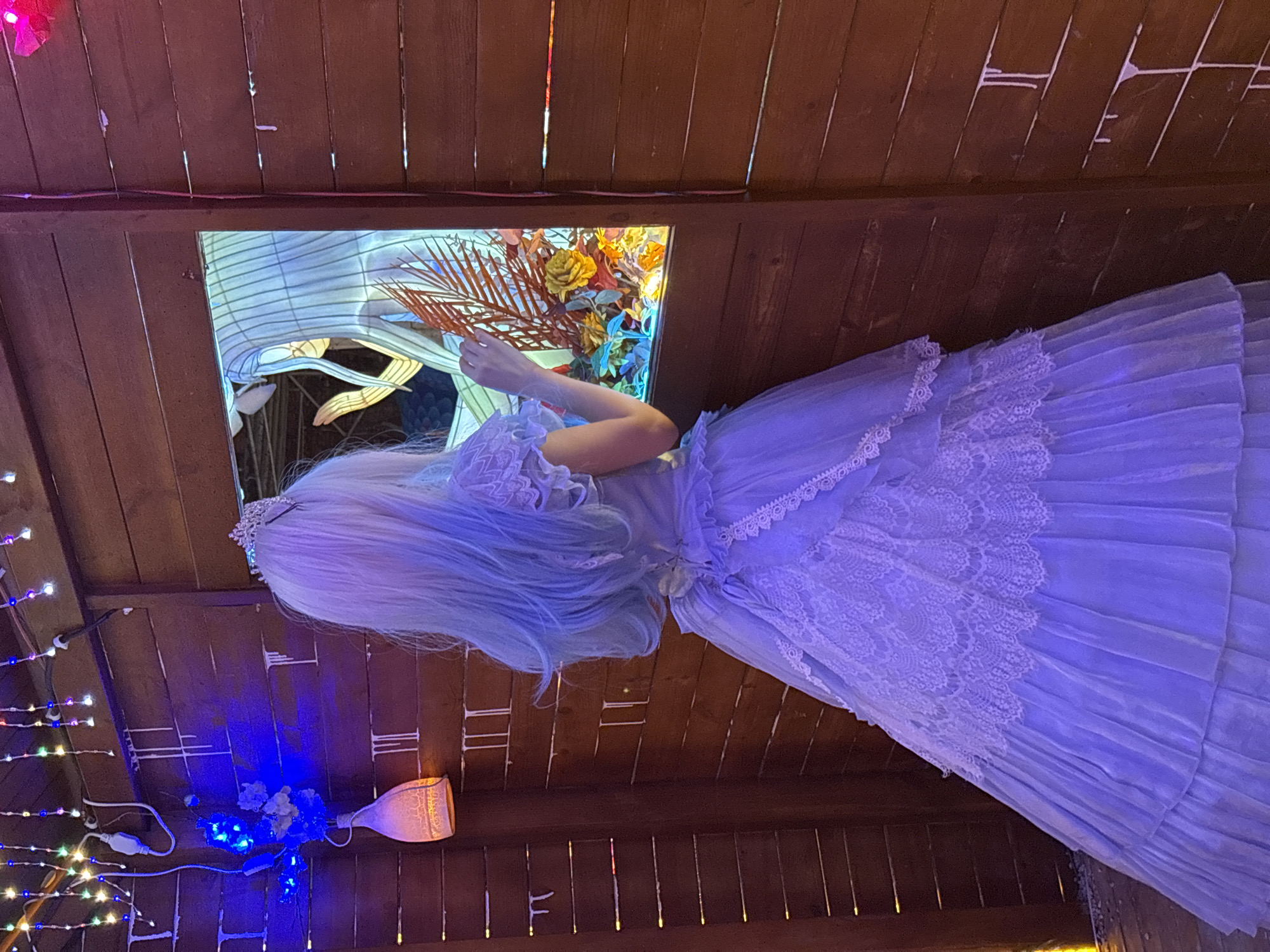 A person wearing a light purple, floor-length gown and a tiara stands facing away from the camera, looking into a window. Their hair is long and appears to be dyed in a gradient of white to light blue. The gown features lace detailing on the bodice and skirt, with multiple layers of ruffled fabric. The person's right hand is extended towards the window.
The scene takes place outdoors, with a wooden structure forming the backdrop. The structure has horizontal planks and appears to be a small booth or display. String lights in various colors are visible hanging from the top of the structure, and blue lights illuminate the left side of the scene. Inside the window, a lantern-like display features stylized figures and floral arrangements, illuminated from within. The ground beneath the person appears to be made of wood or a similar material. The overall impression is a festive or decorative setting, possibly part of a market or an exhibition. The location is identified as Baqiao, China.