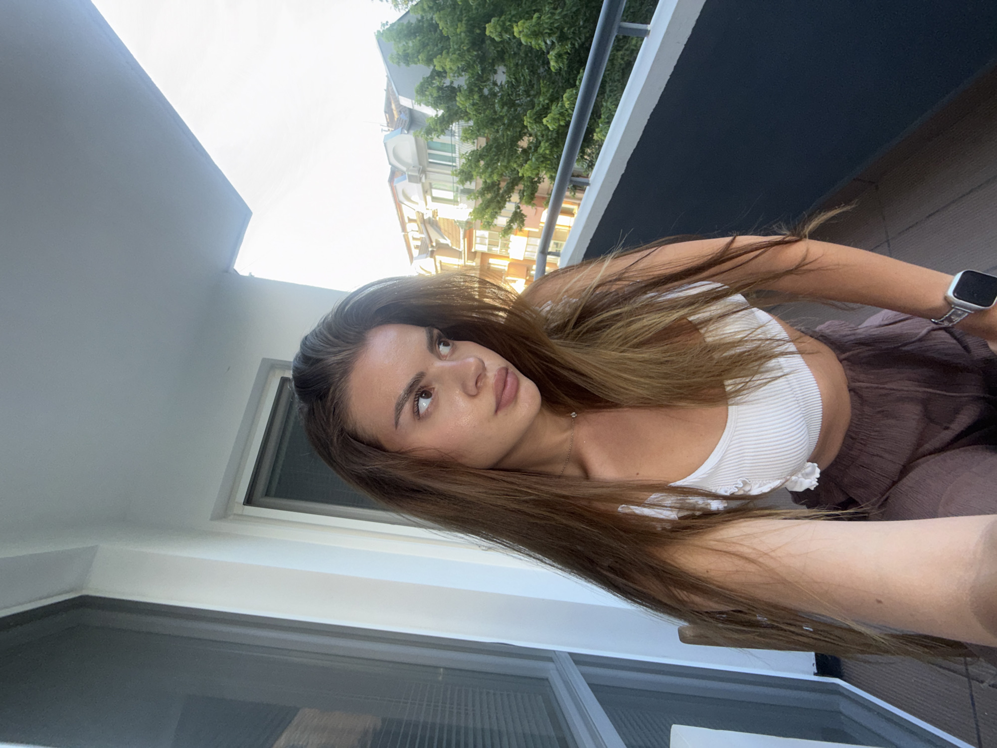 A young woman with long, straight brown hair, parted slightly off-center, is captured in a medium shot on a balcony. She is looking intently to her right, away from the camera, with a pensive expression. Her lips are full, and her eyebrows are well-defined. She is wearing a white ribbed crop top with ruffled detailing on the straps and loose-fitting, crinkled brown pants. A delicate necklace with a small, singular pendant is visible around her neck. On her left wrist, she wears a silver Apple Watch with a dark band.
The scene is set on a balcony in Plovdiv, Bulgaria. To the left, a large window or glass door with internal blinds is visible within a white frame. Above, a white ceiling overhangs the balcony. The balcony floor is tiled in a light, neutral color. To the right, a dark grey balcony railing with a lighter grey handrail spans the width of the frame. Beyond the railing, an urban landscape unfolds under what appears to be late afternoon or early evening light, casting soft shadows. Multiple multi-story buildings with pitched roofs are visible, exhibiting a style consistent with European architecture. One prominent building directly behind the woman's right shoulder features a cream-colored facade, brown roof tiles, and distinct dormer windows. Green foliage from trees is interspersed among the buildings, indicating a leafy urban environment.